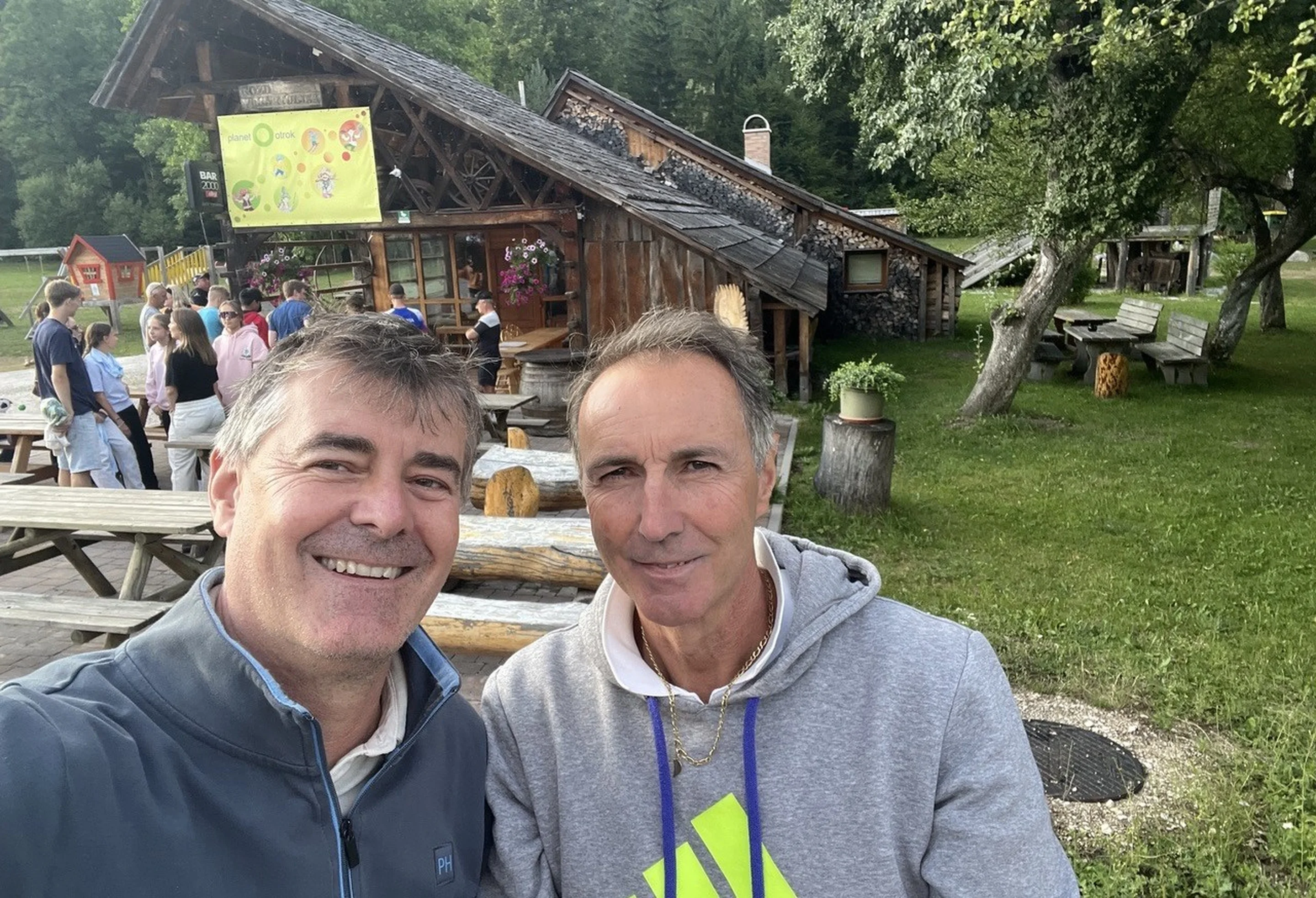 Two men smiling outdoors with a rustic wooden building in the background and people gathered around picnic tables.