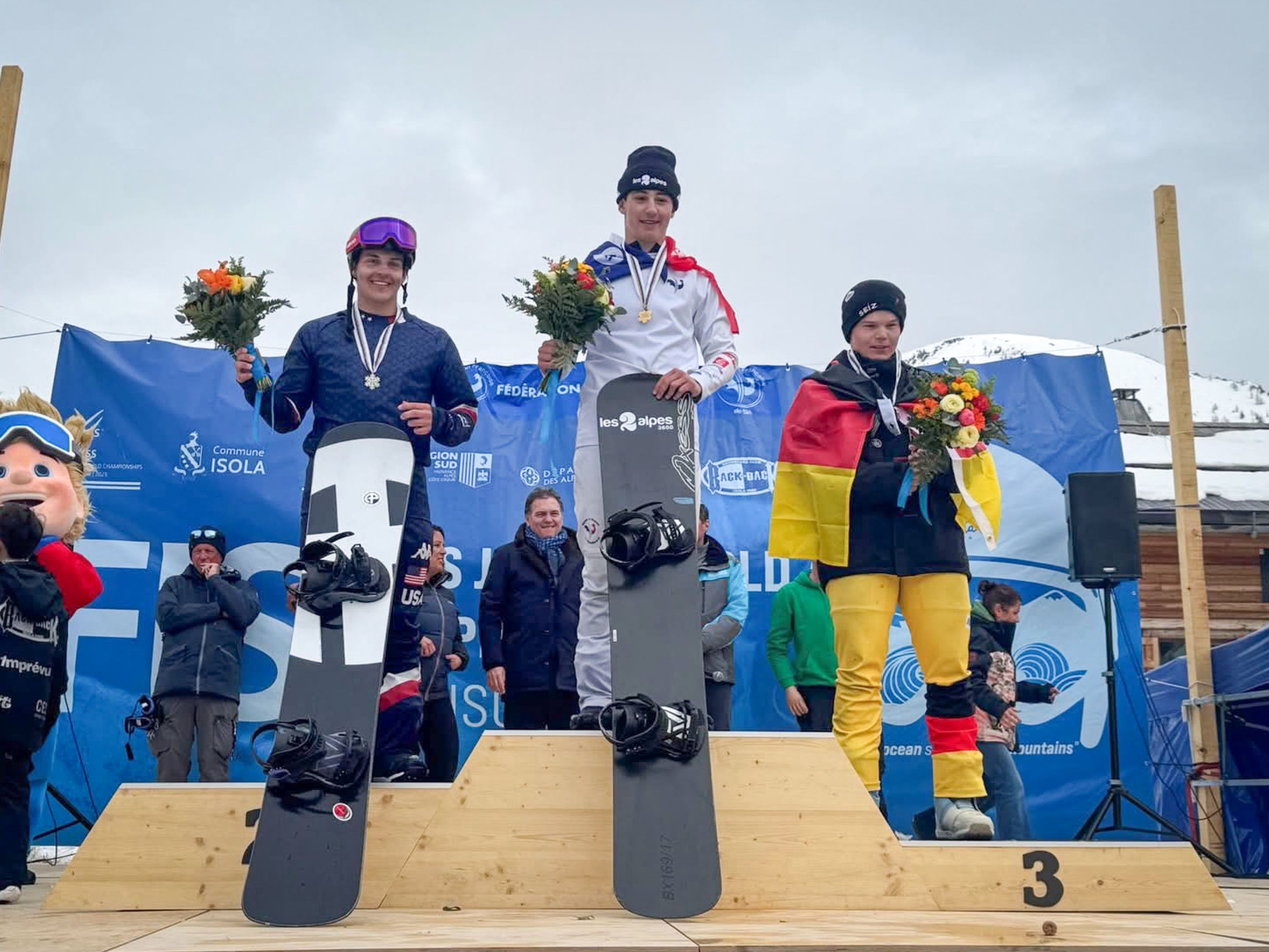 Three snowboarders stand on a wooden podium wearing medals and holding flowers at the 2025 Junior World Championships, with Jonas Chollet in white in the middle