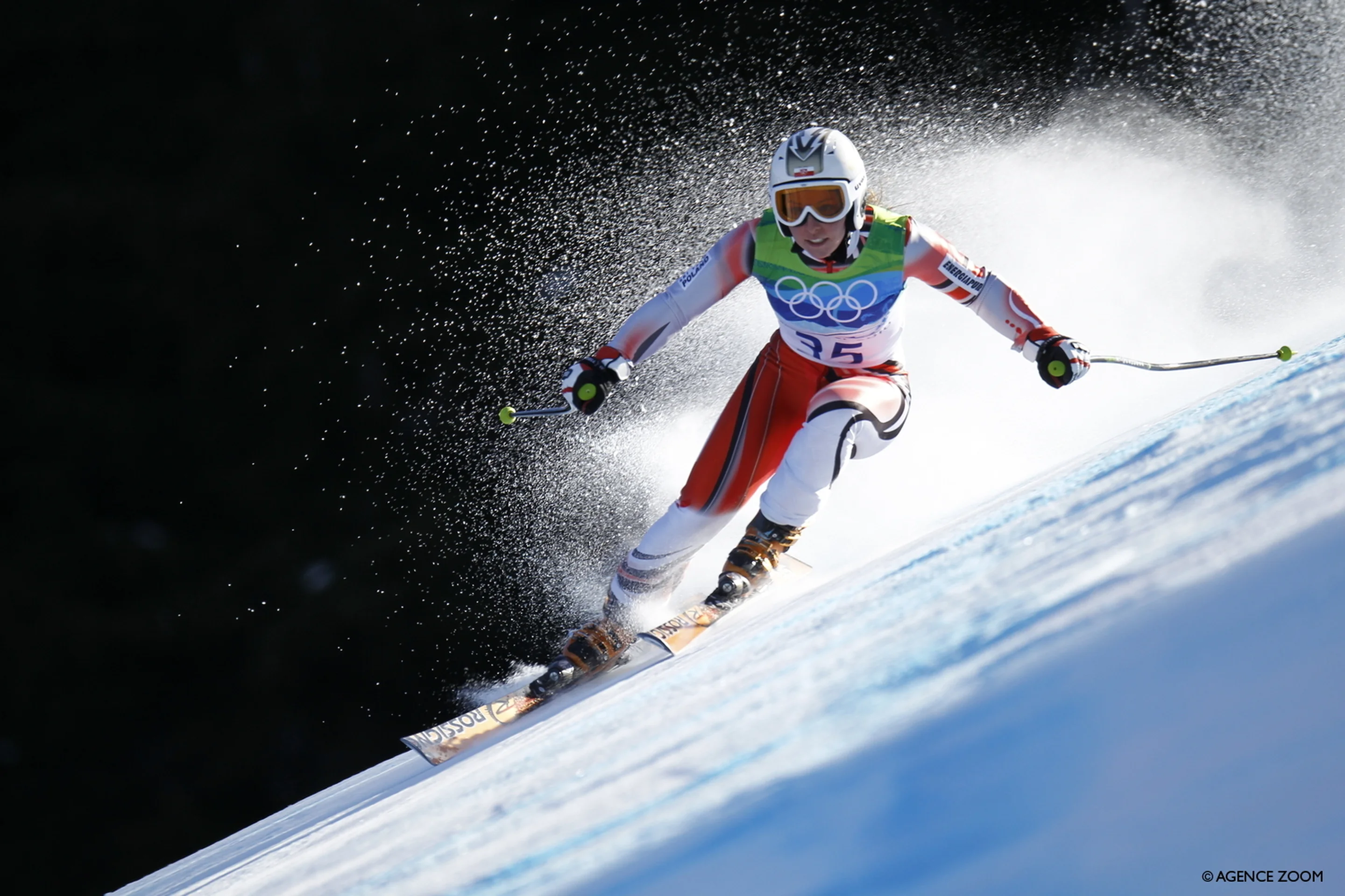 A skier in Olympic gear navigates a downhill slope, snow spraying dramatically, against a backdrop of shadowed terrain.