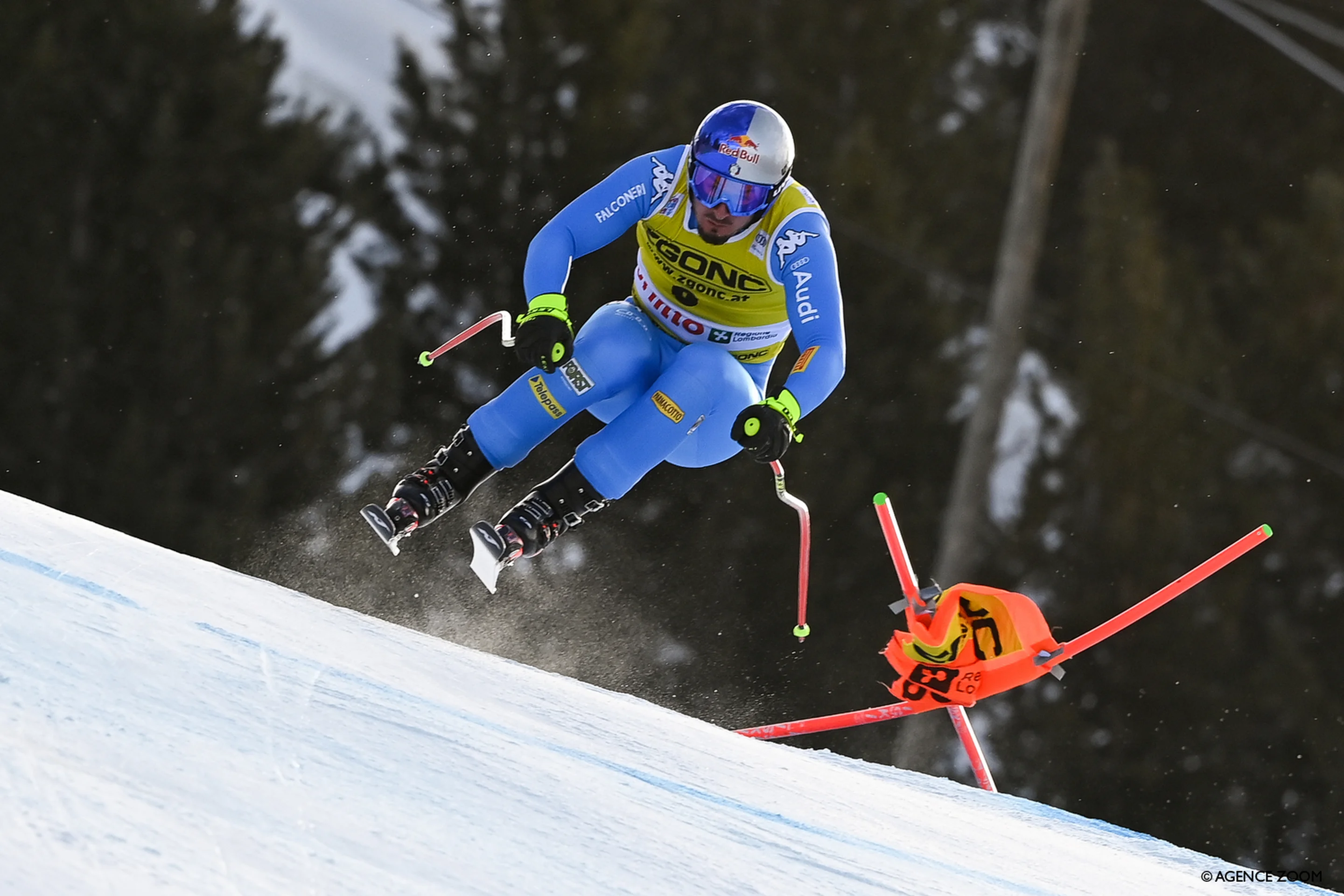 A skier in blue gear, mid-air during a downhill race, with a bent ski pole and a marker in the snow-covered mountain setting.