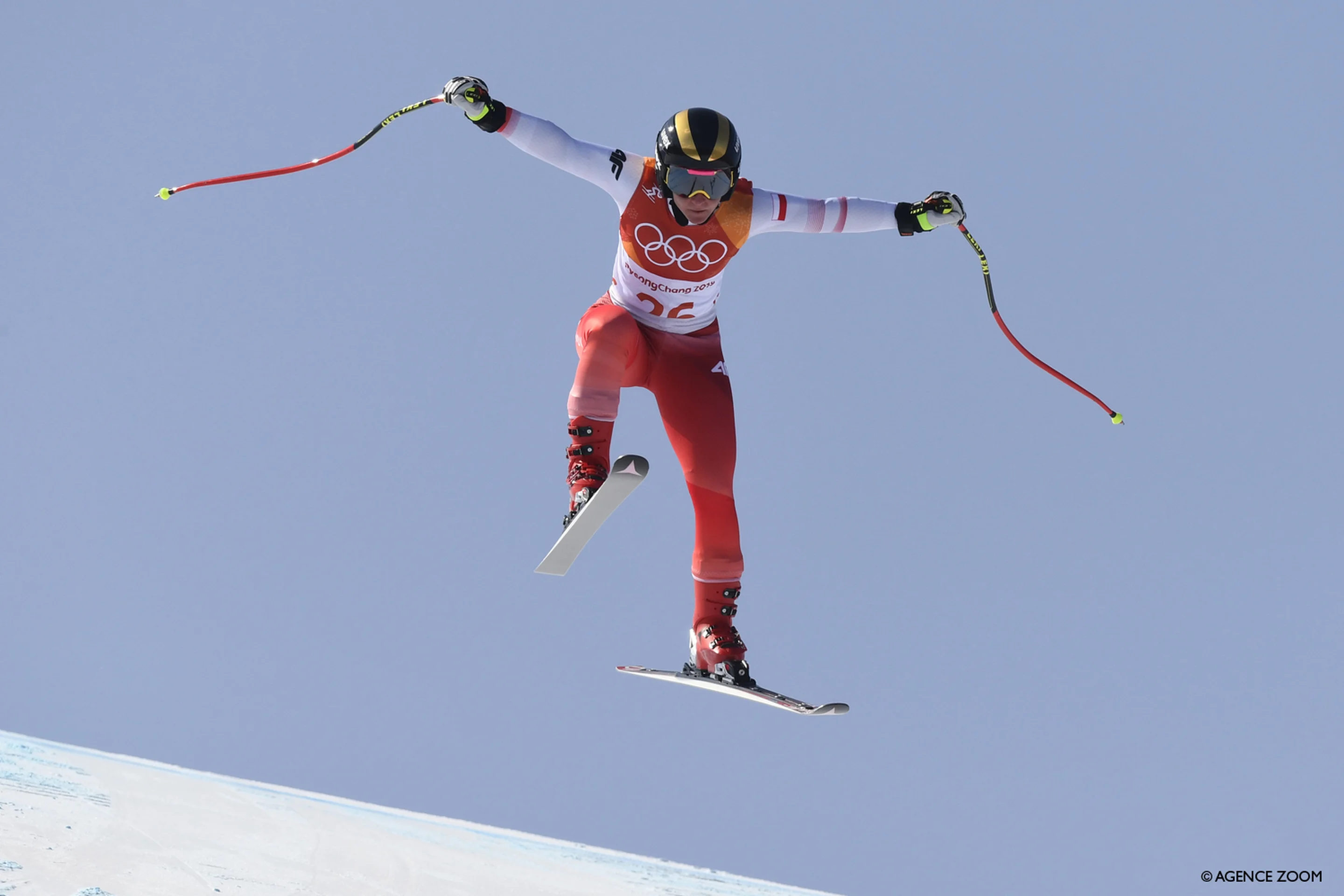 Skier in mid-air, wearing red and white gear with Olympic rings visible, skiing downhill against a clear blue sky.