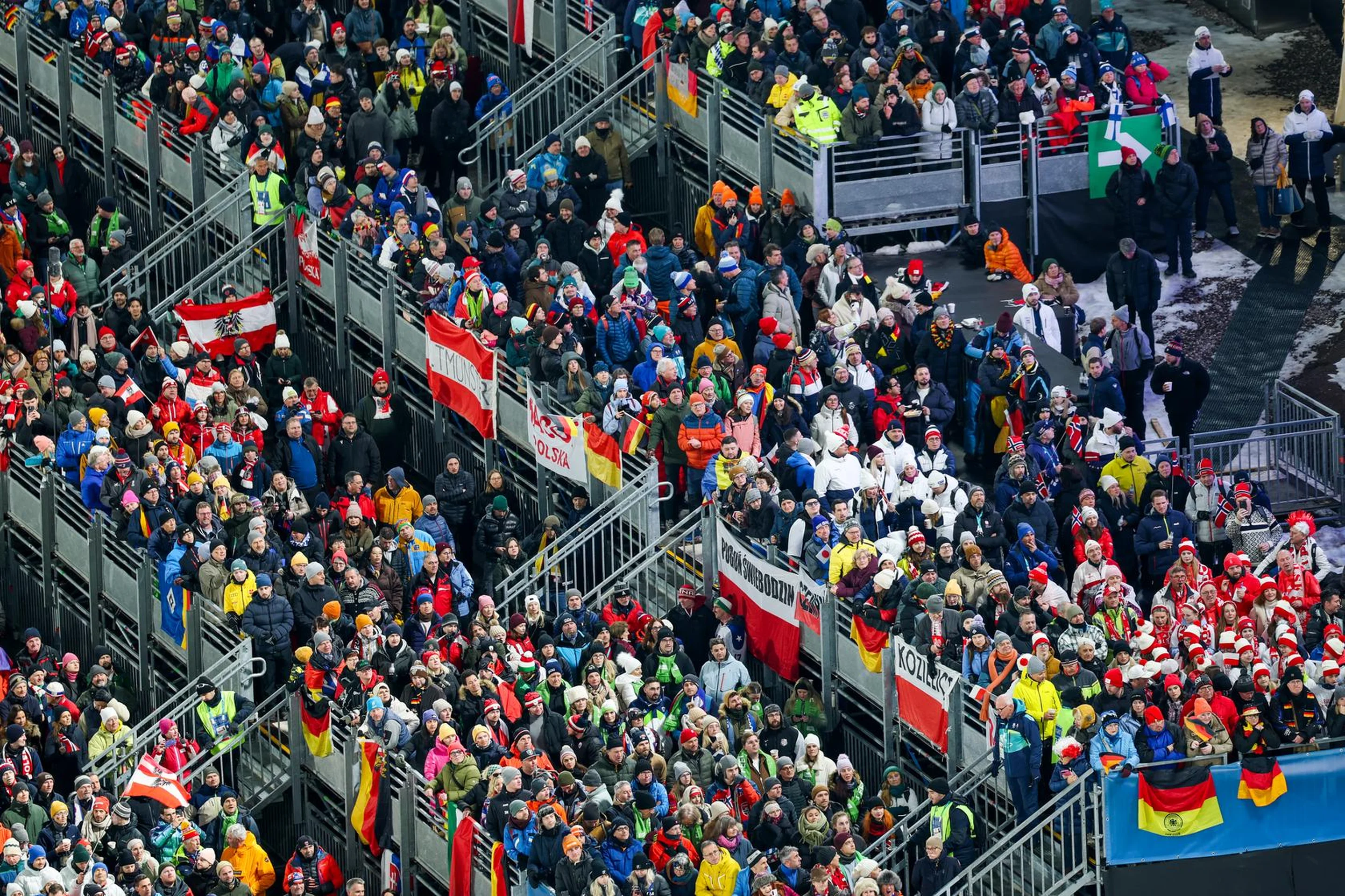 A large crowd of people in colorful clothing gathers in stadium stands, holding various national flags, creating a vibrant and lively scene.