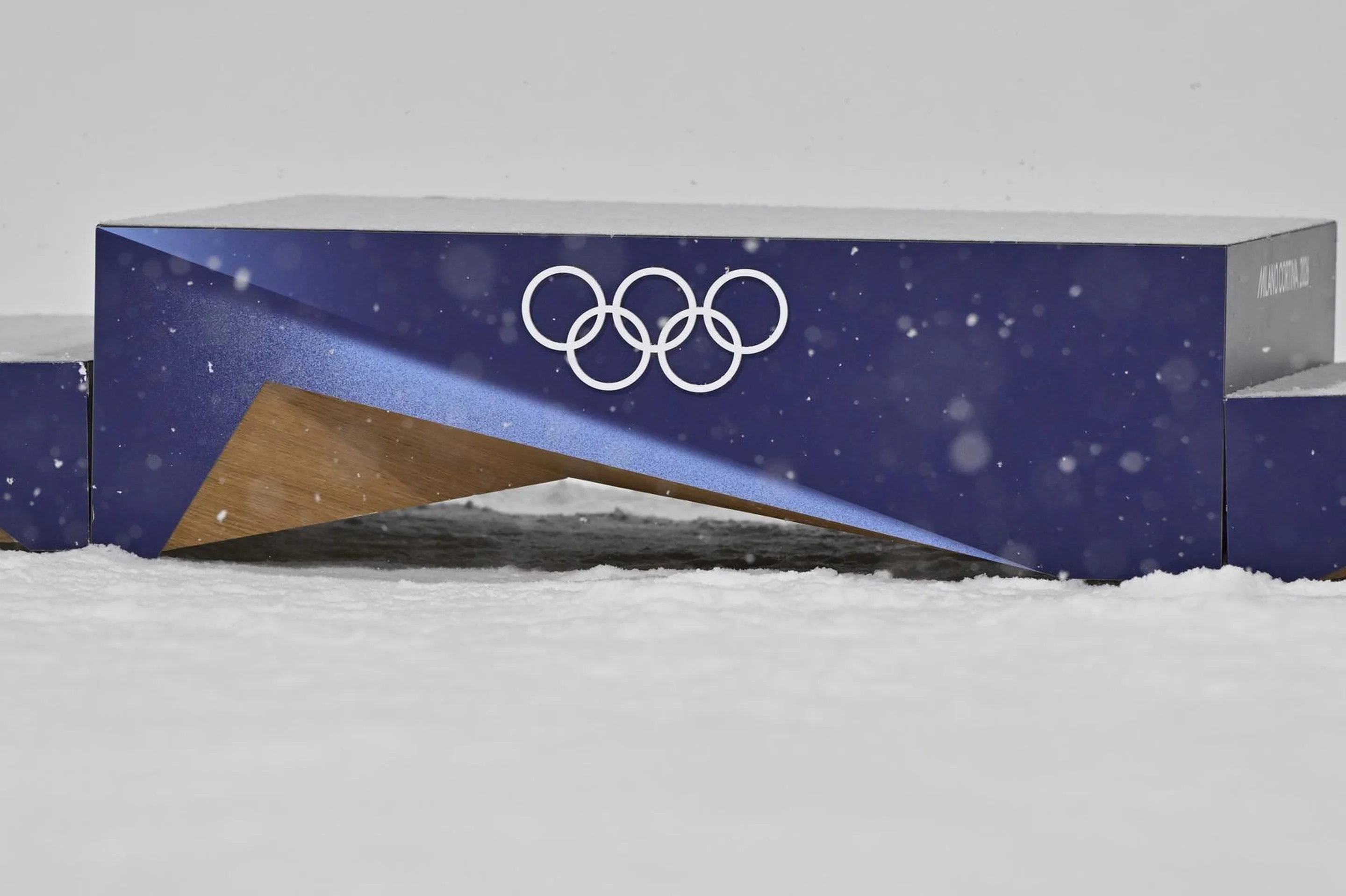 Olympic podium with three levels, featuring the Olympic rings on a blue background, surrounded by snow.
