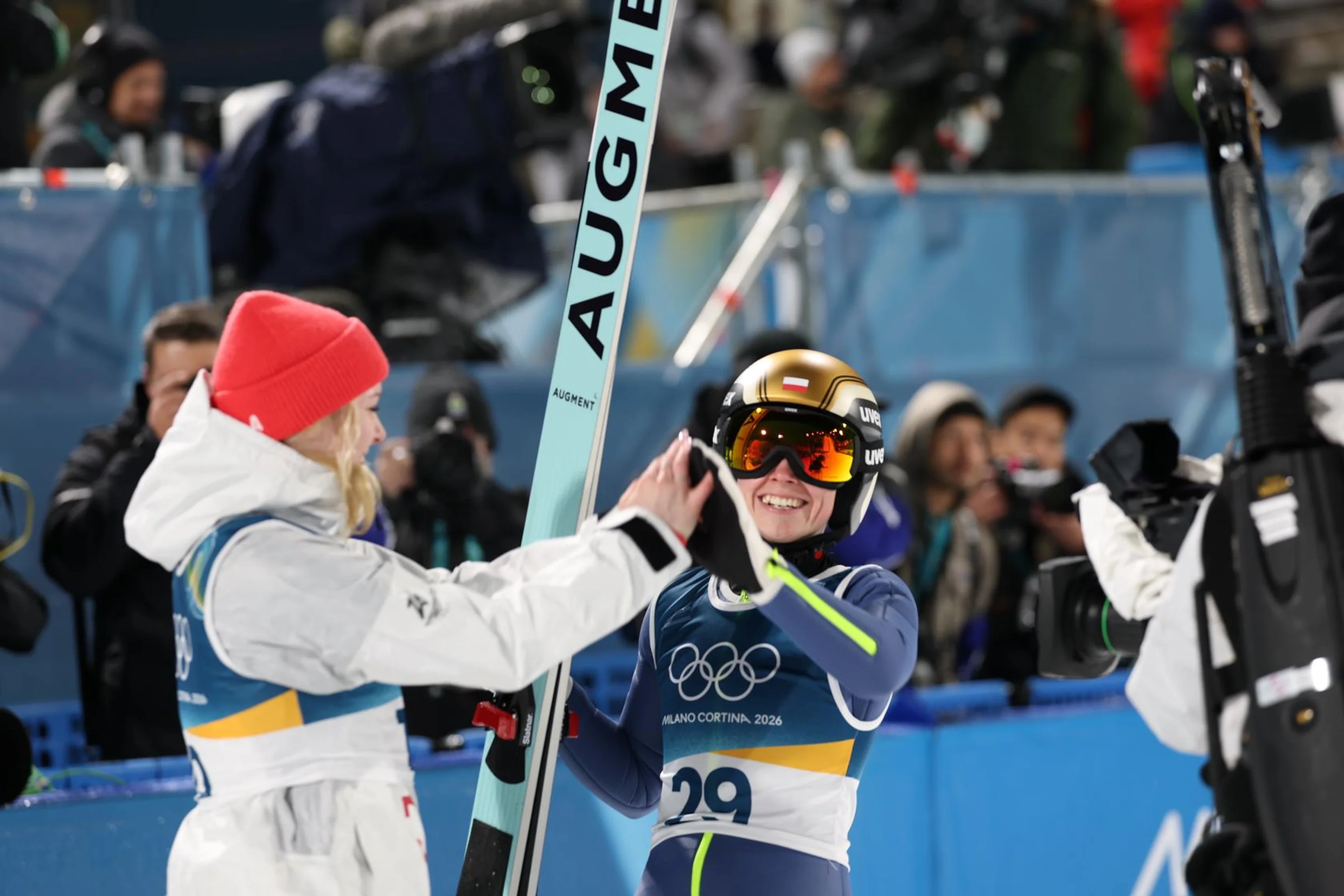 Two athletes high-five at a winter sports event, surrounded by photographers. One wears a red hat; the other holds a ski with "AUGMENT" on it.