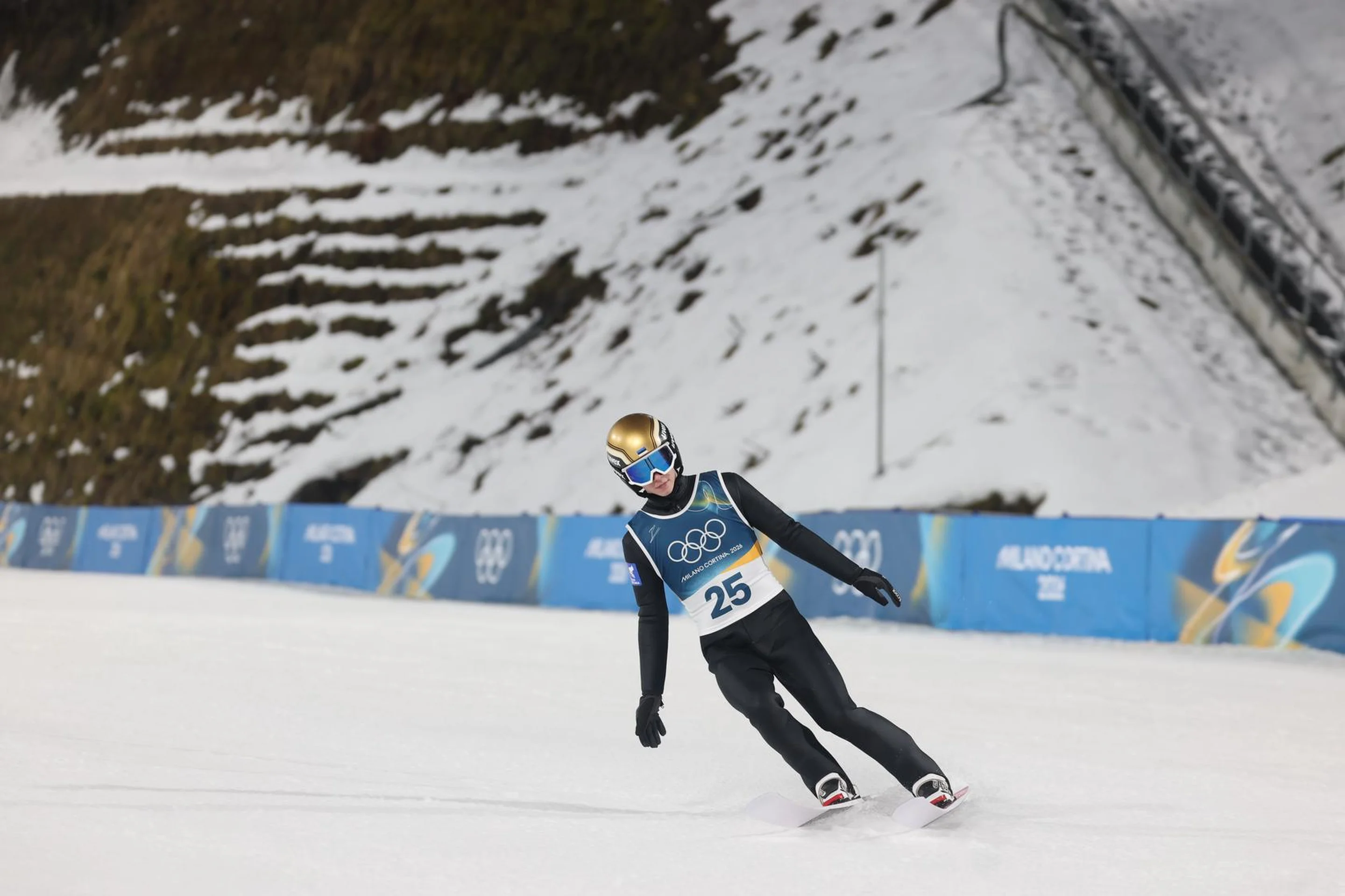 A skier wearing a blue bib and helmet glides on a snowy slope at a winter sports event, with banners and snow-covered steps in the background.