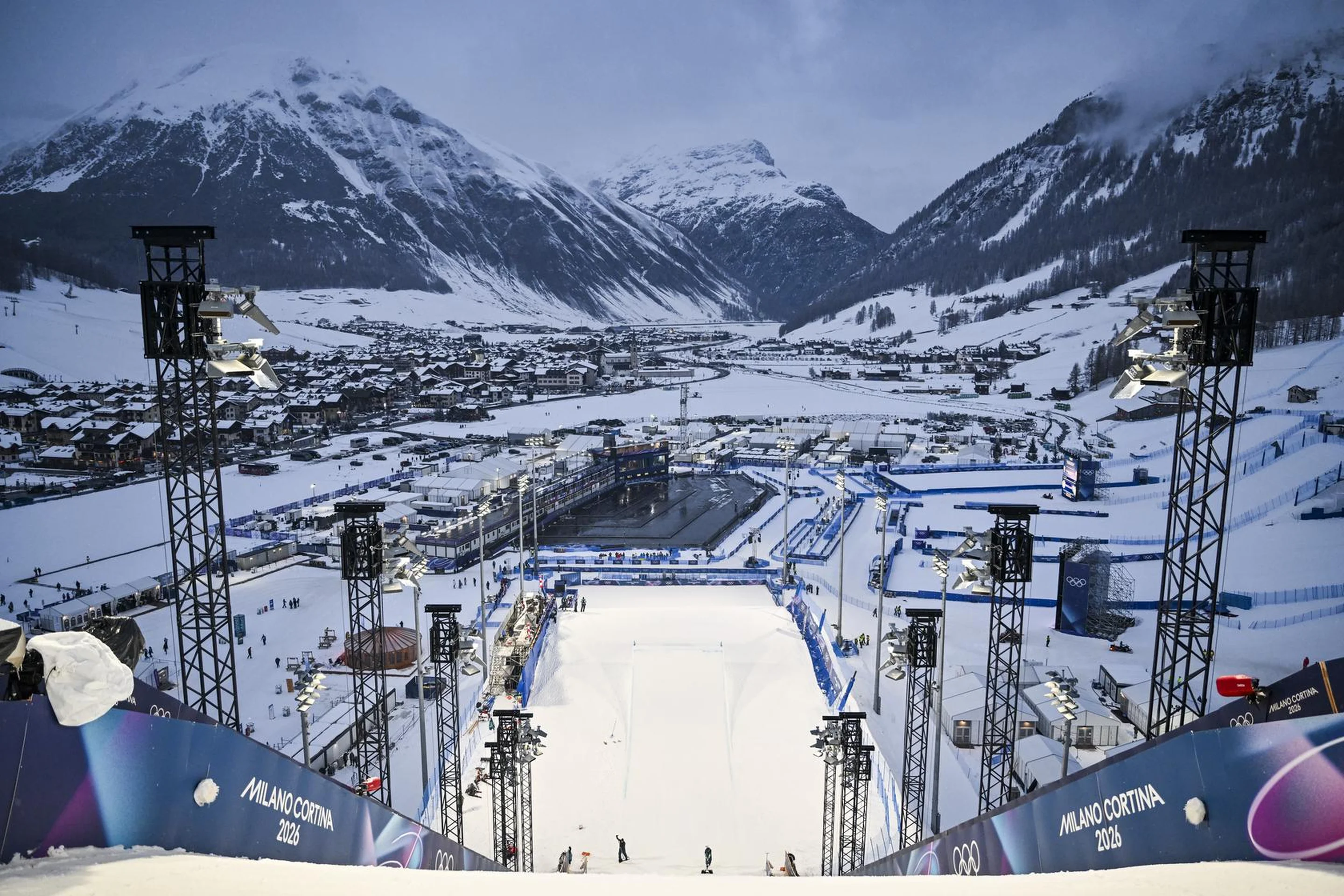 View from a ski jumping hill overlooking a snowy valley with mountains, lit by tall spotlights, and a village in the background.