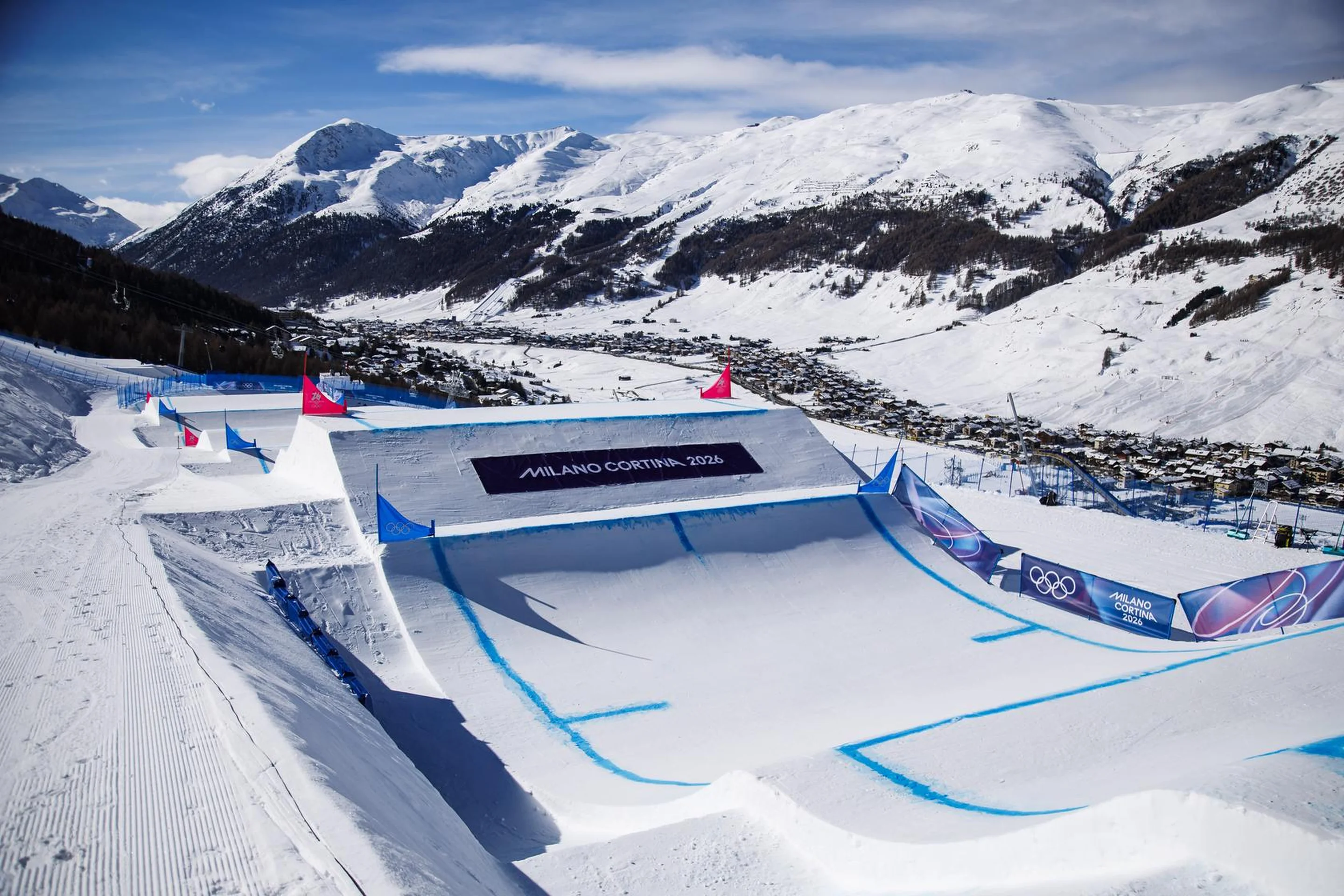 Snowy Olympic ski slope with a large ramp, blue markings, and distant mountains under a clear sky. Banners display "Milano Cortina 2026."