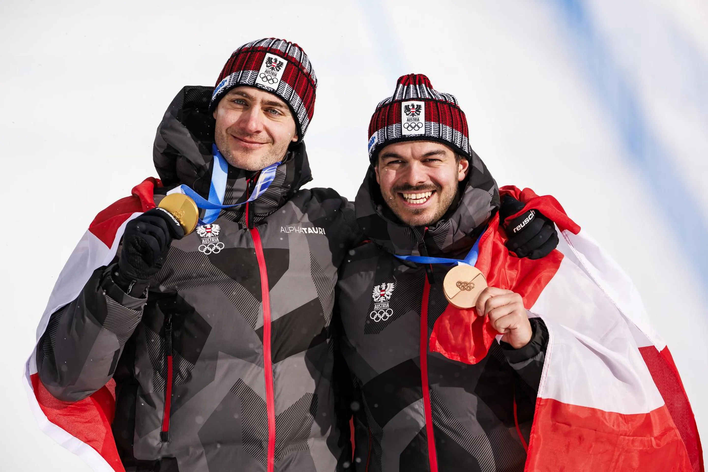 Two athletes in winter gear celebrate with gold and bronze medals, wearing beanies, and wrapped in flags against a snowy background.