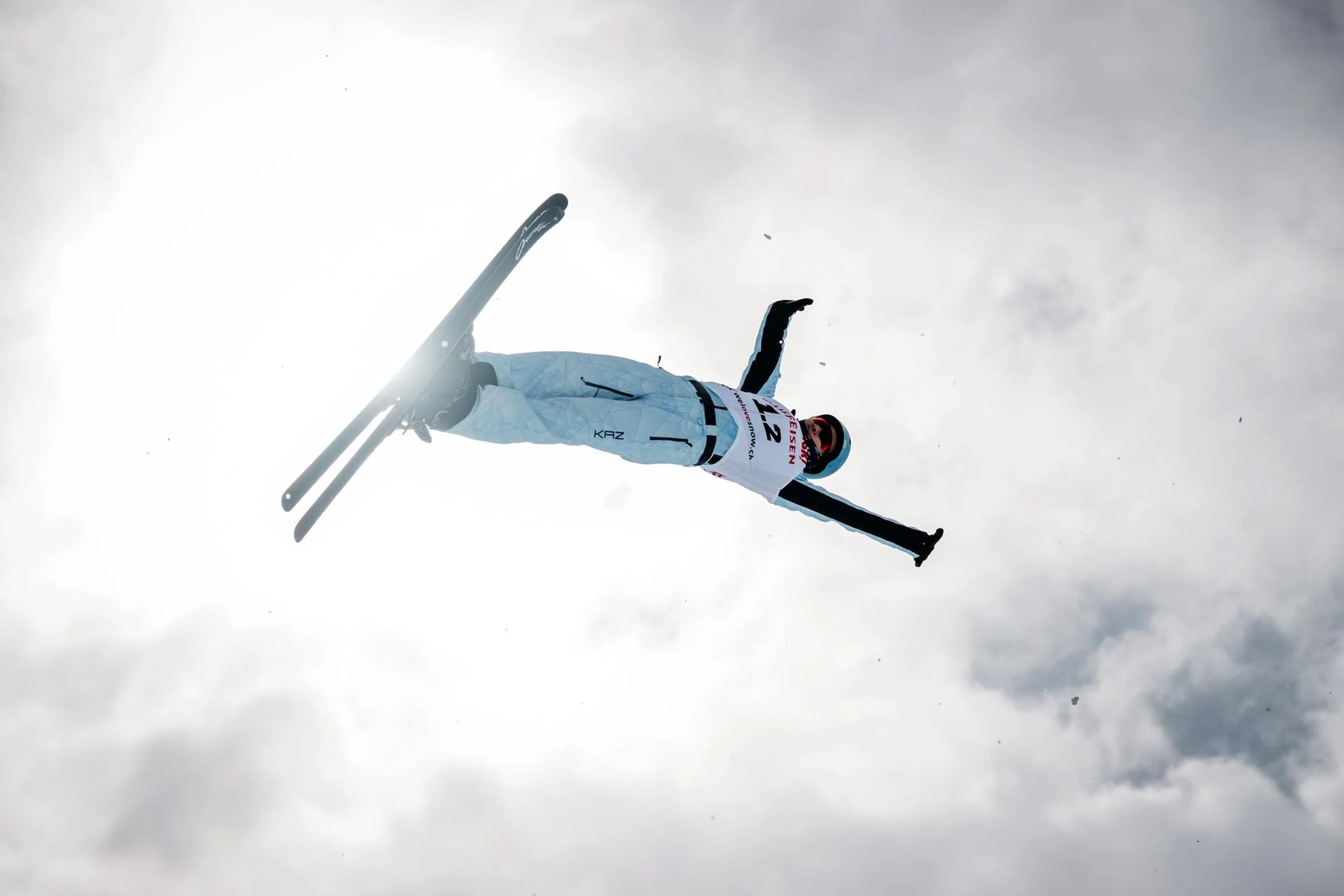 A skier performs an aerial trick against a cloudy sky, arms outstretched, with sunlight gleaming in the background.