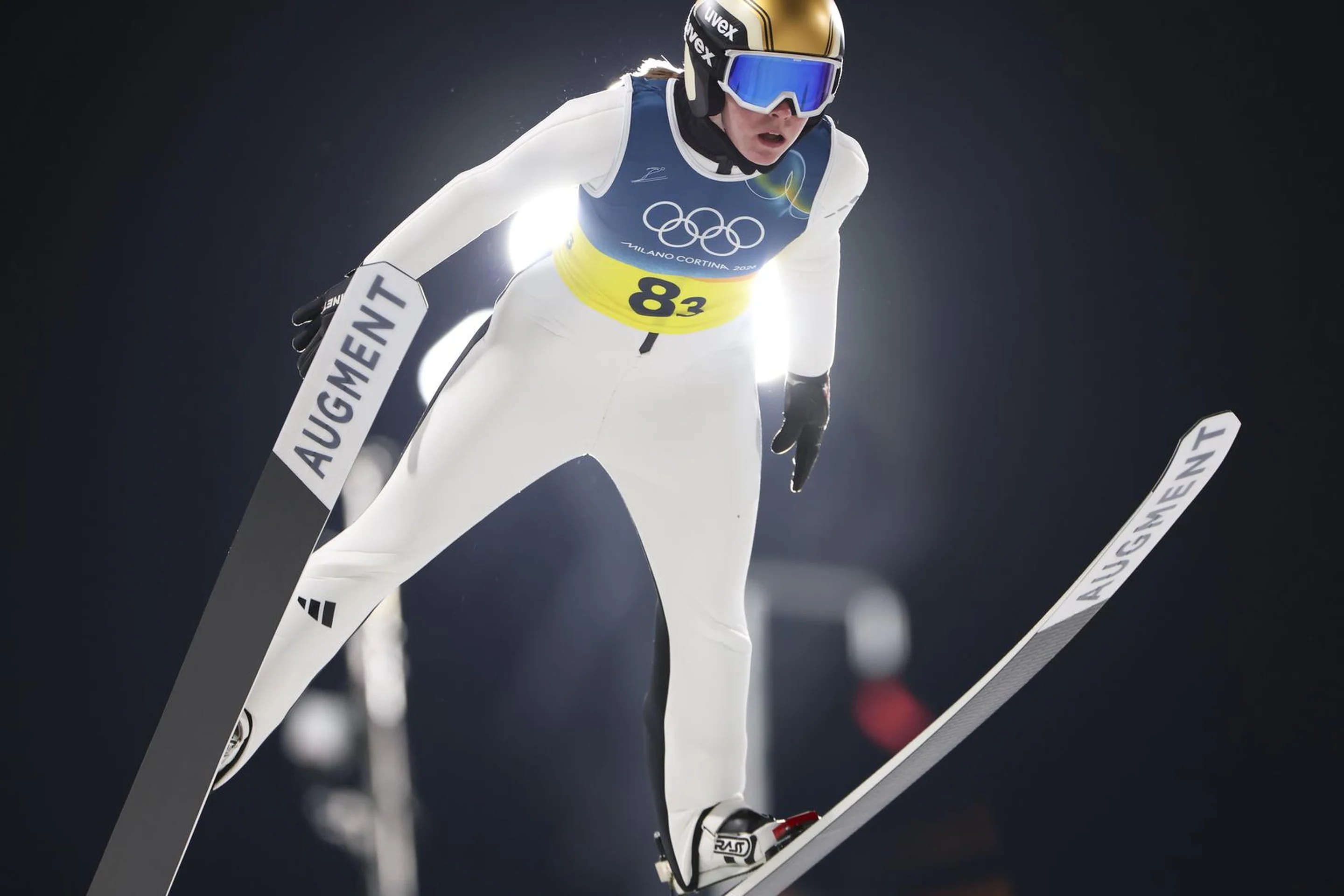 Ski jumper in mid-air wearing a white suit and helmet with a gold visor, dark background highlighting the dynamic pose.