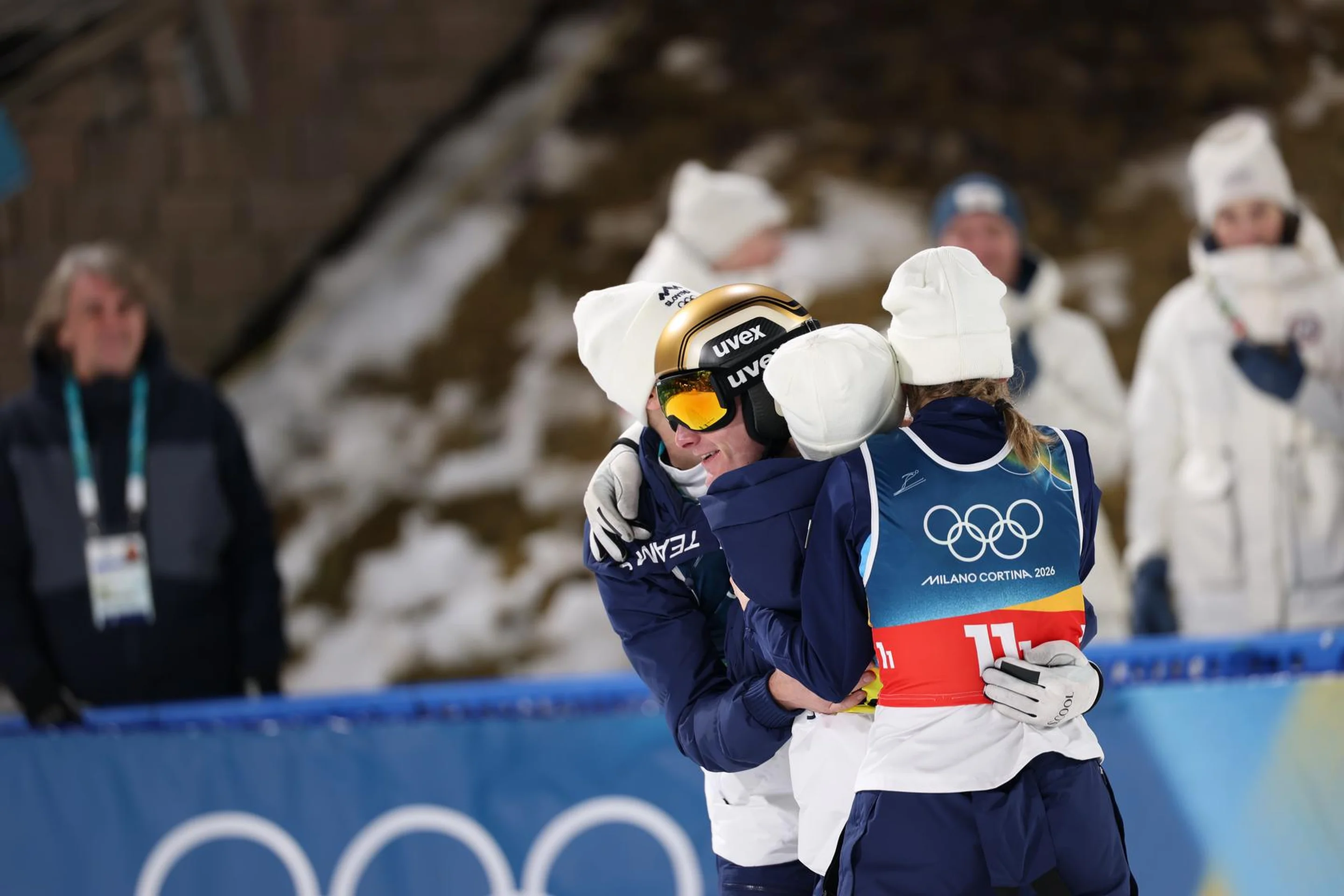 Three athletes in winter gear embrace on a snowy slope with Olympic branding in the background, surrounded by spectators.