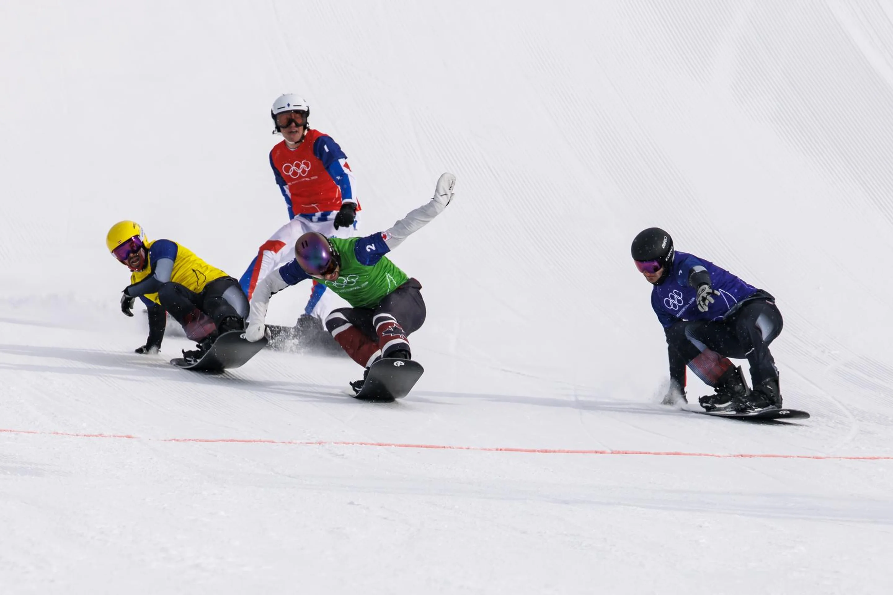 Four snowboarders racing downhill on a snowy slope, wearing colorful gear and helmets, with an Olympic logo visible in the background.