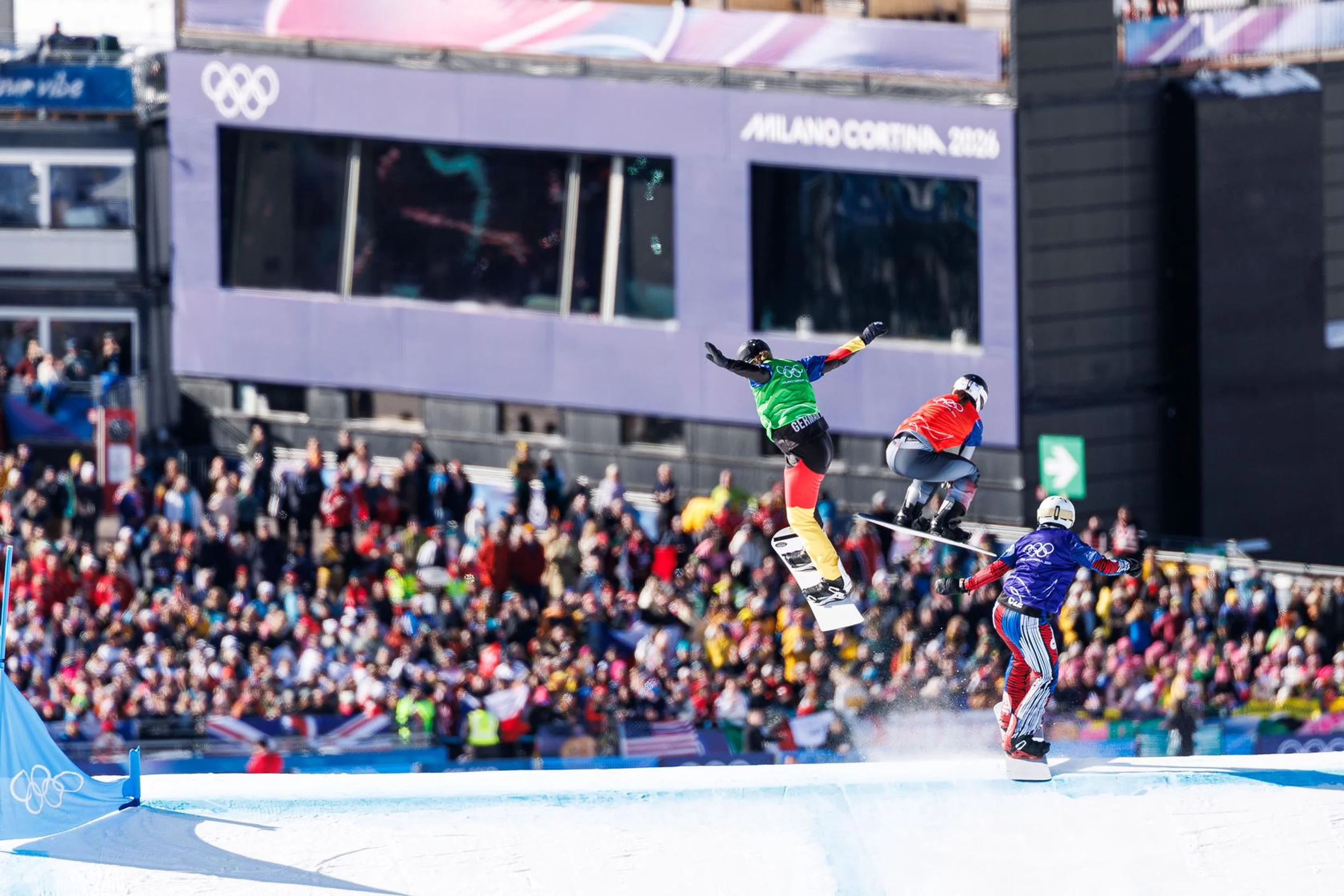 Snowboarders in mid-air during a competitive race at the Olympics, with a large crowd and event banners in the background.