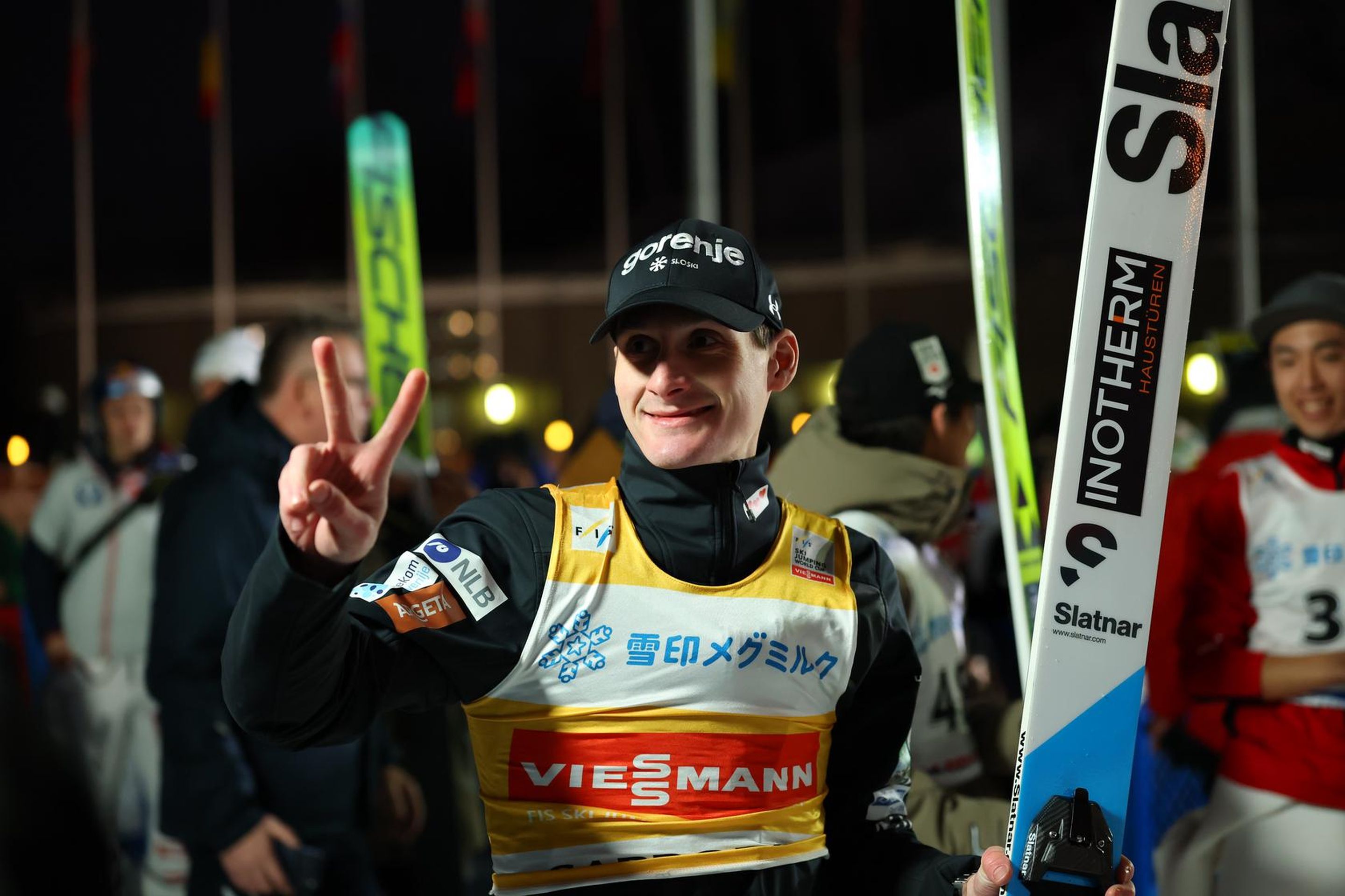 A ski jumper in a yellow bib holds skis and flashes a peace sign, smiling at an outdoor event with people and flags in the background.