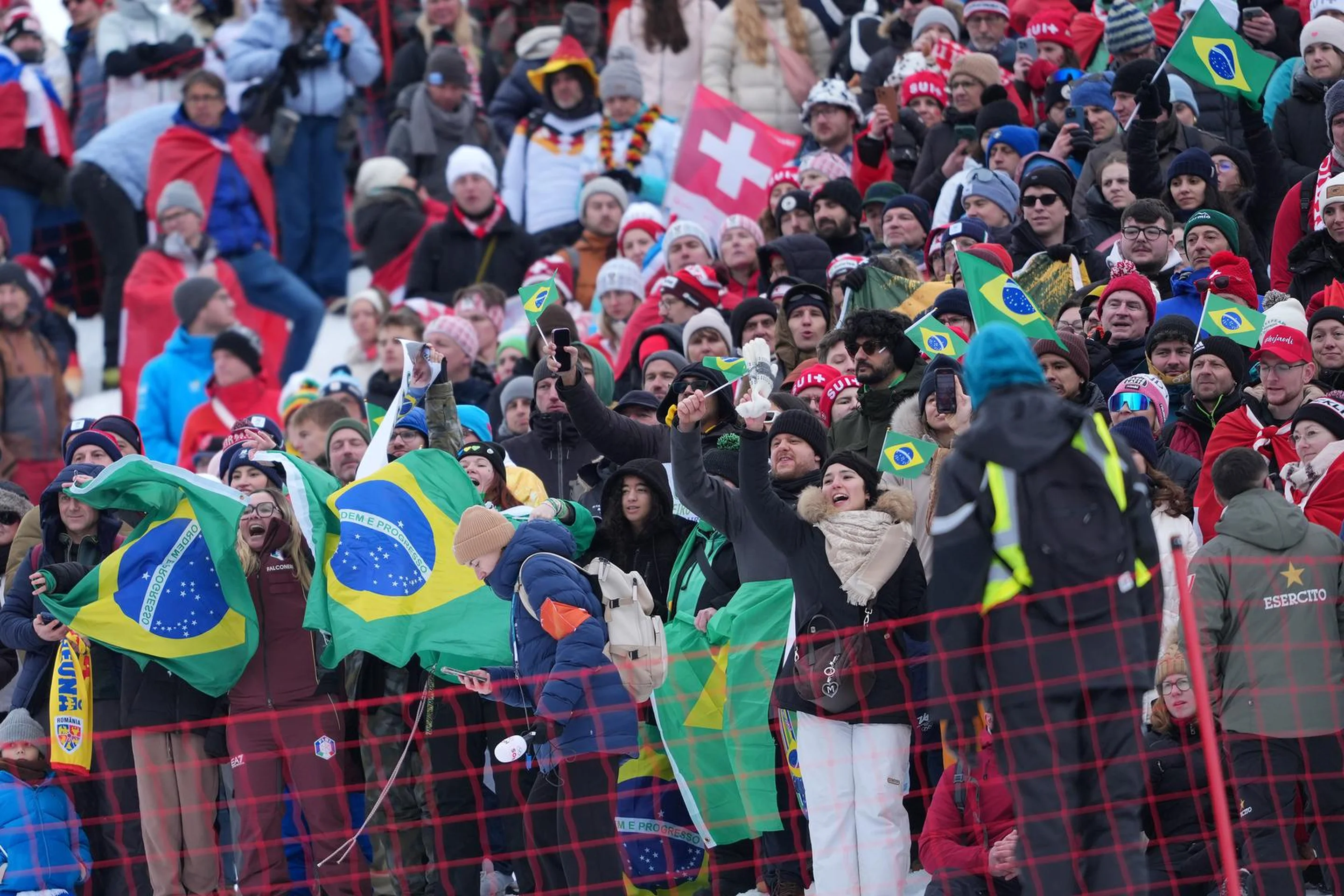 A lively crowd at a sports event holds Brazilian flags, with many fans dressed warmly and some wearing colorful outfits and accessories.