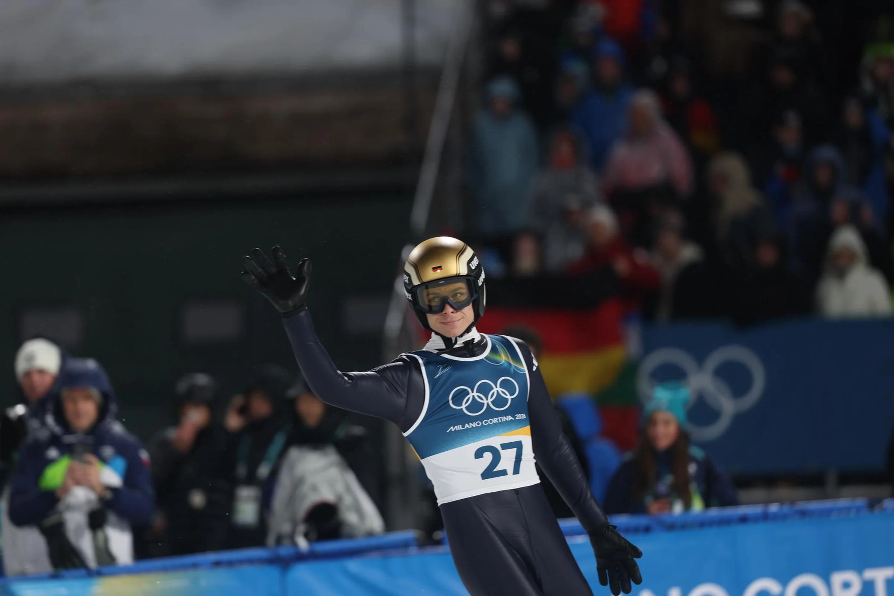 A ski jumper in a helmet and goggles waves to the crowd, with Olympic rings and spectators in the background during a winter event.