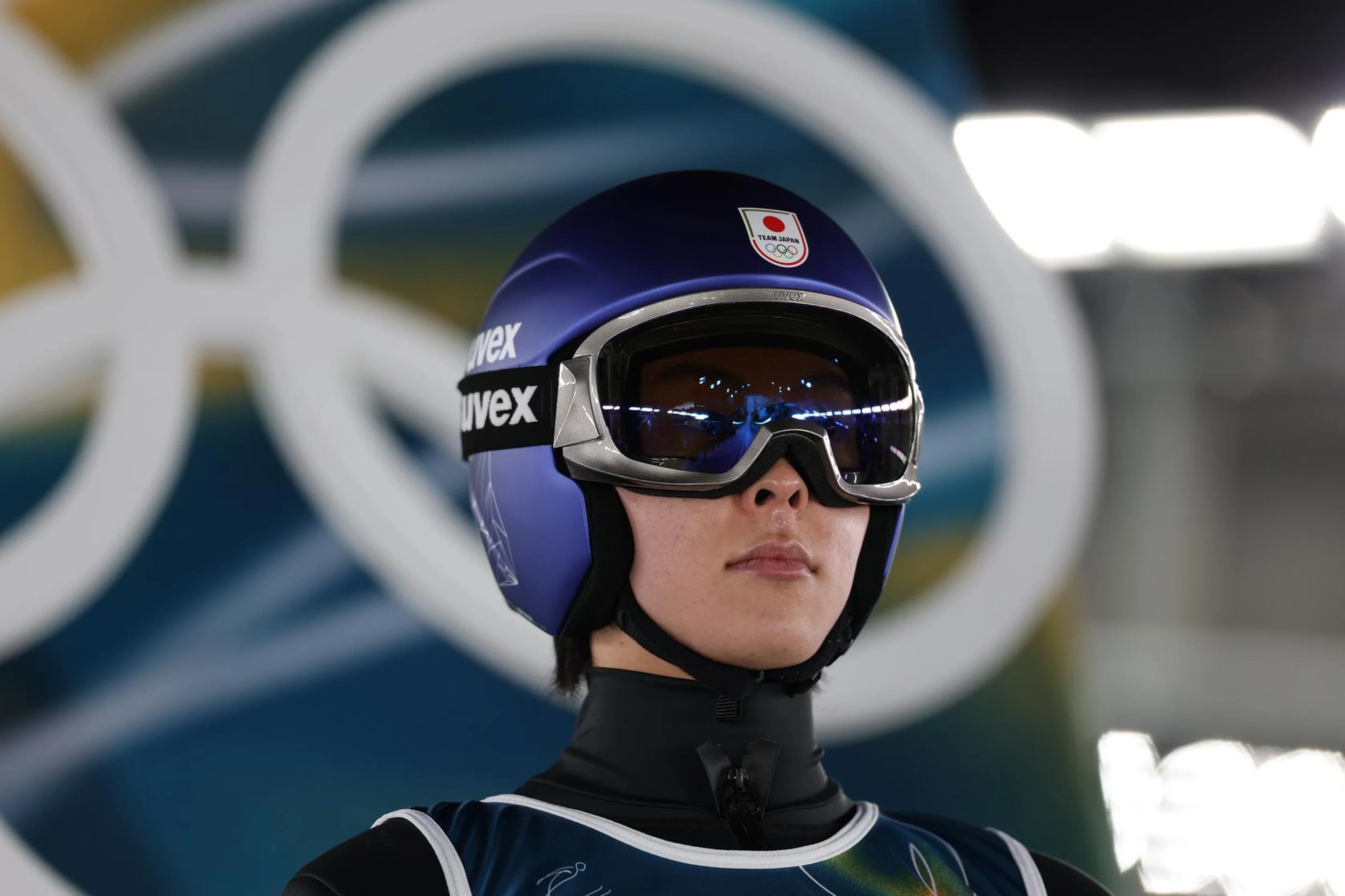 Athlete in ski gear and purple helmet with goggles, stands focused, with Olympic rings blurred in the background.