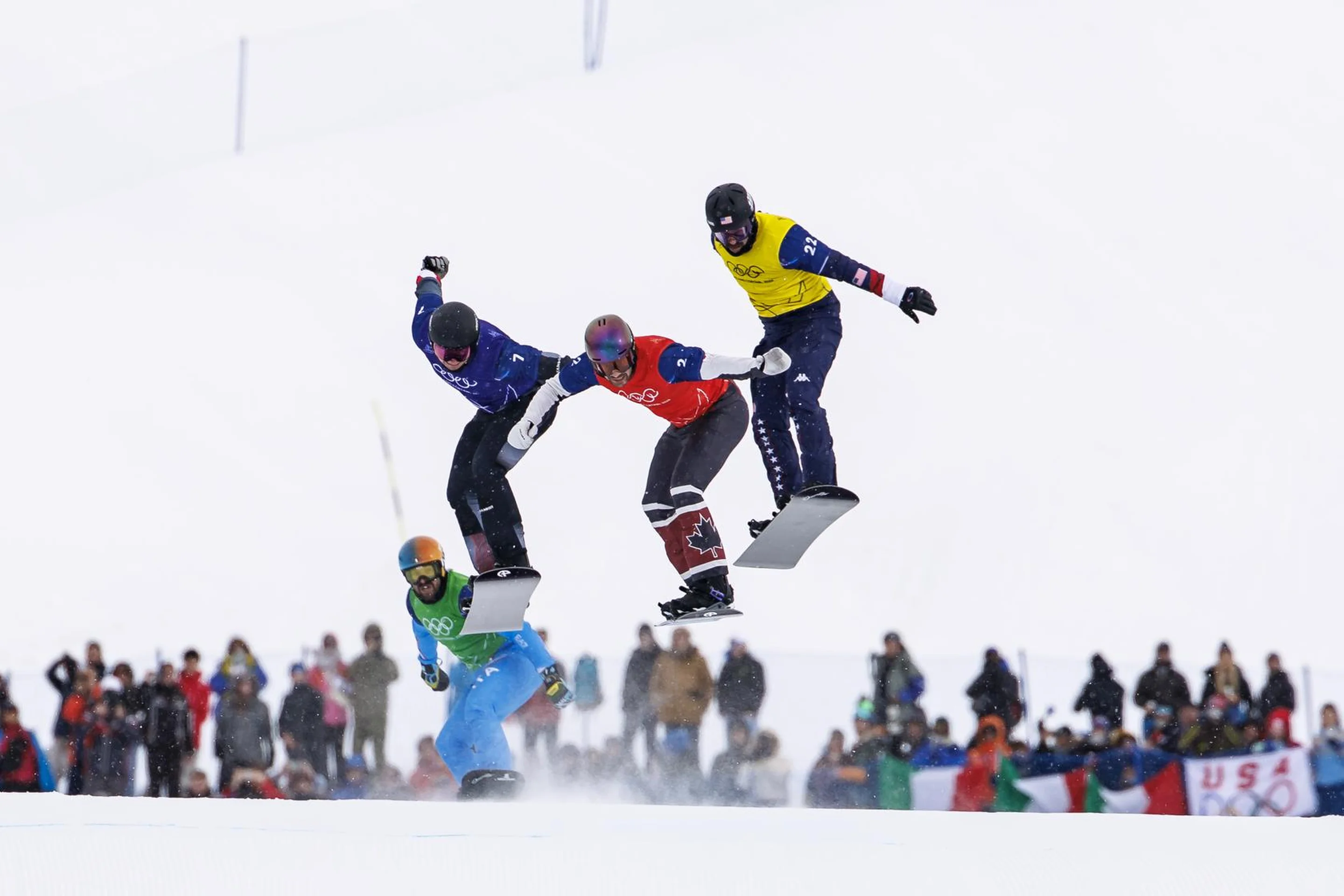 Four snowboarders in mid-air during a race, with spectators in the snowy background, and visible national flags.
