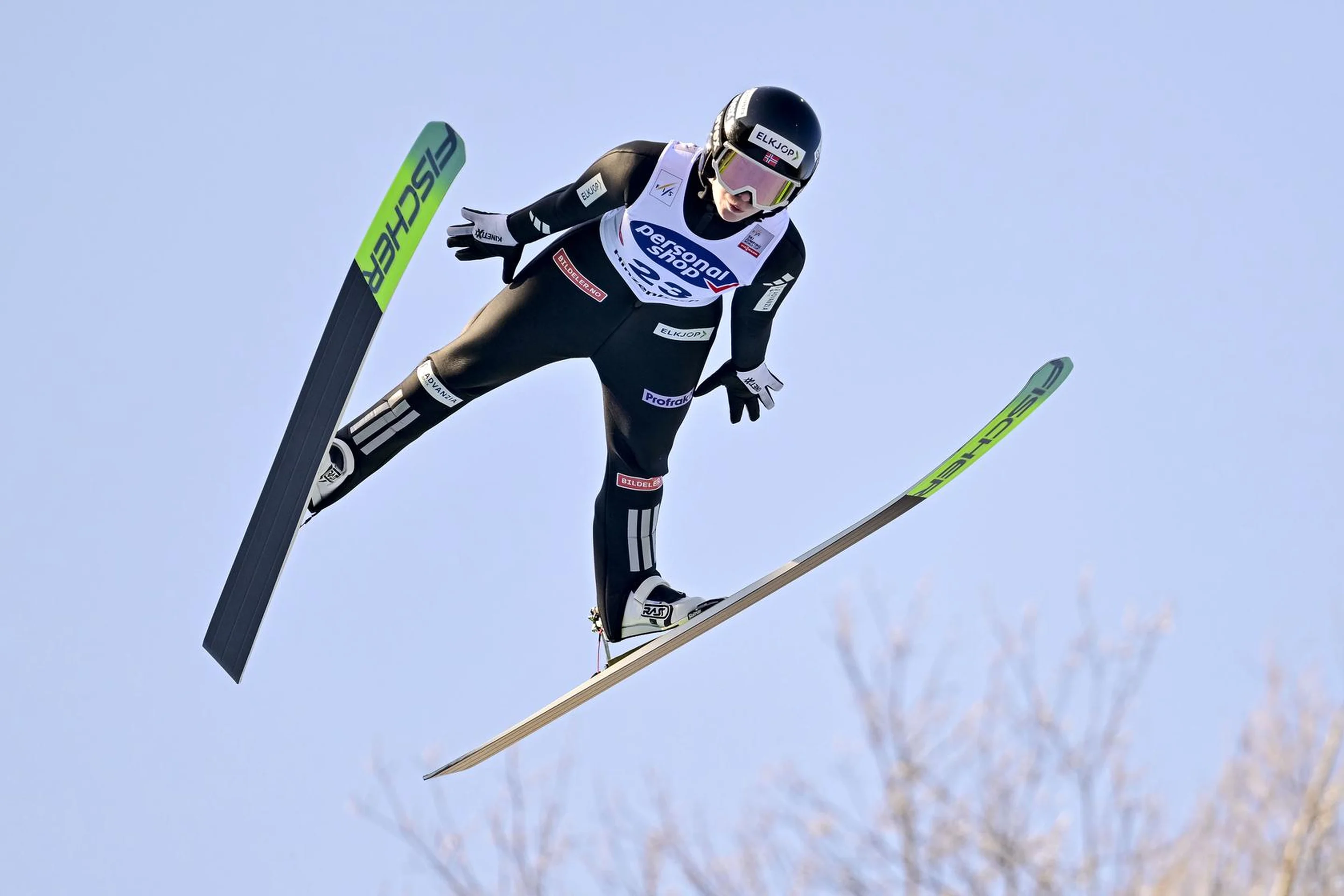 A ski jumper in mid-air wearing a black suit and helmet with green and black skis against a clear blue sky.