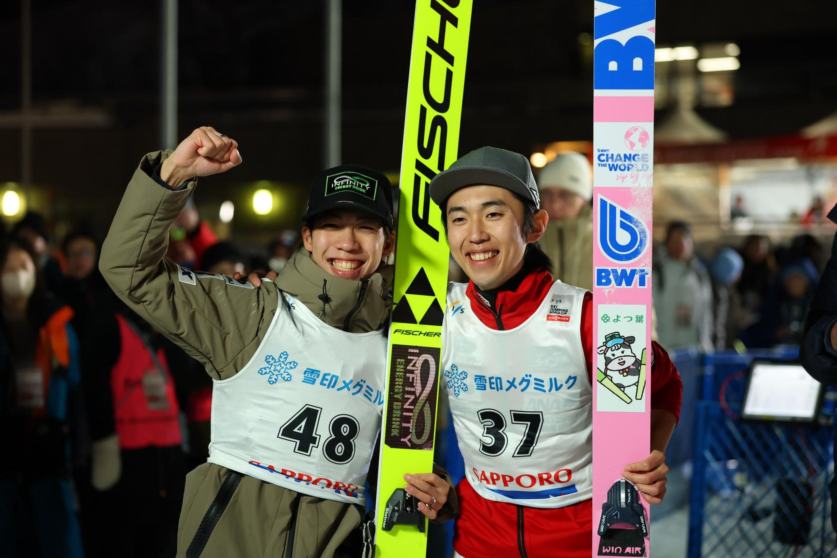 Two ski jumpers celebrating, wearing numbered bibs 48 and 37, with skis and winter gear, in a lively outdoor venue.