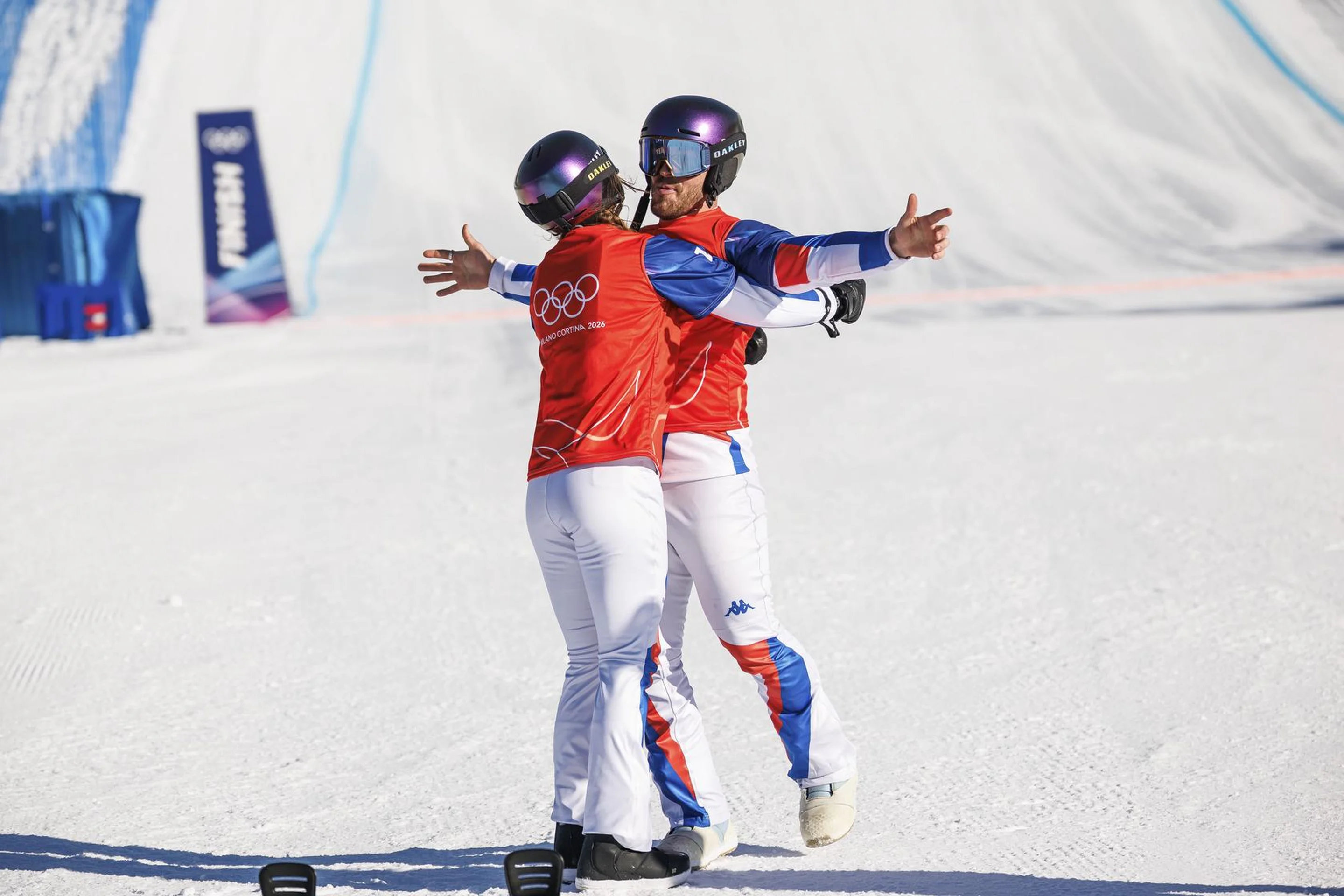 Two snowboarders in red uniforms and helmets embrace joyfully on a snowy slope, celebrating a successful event.