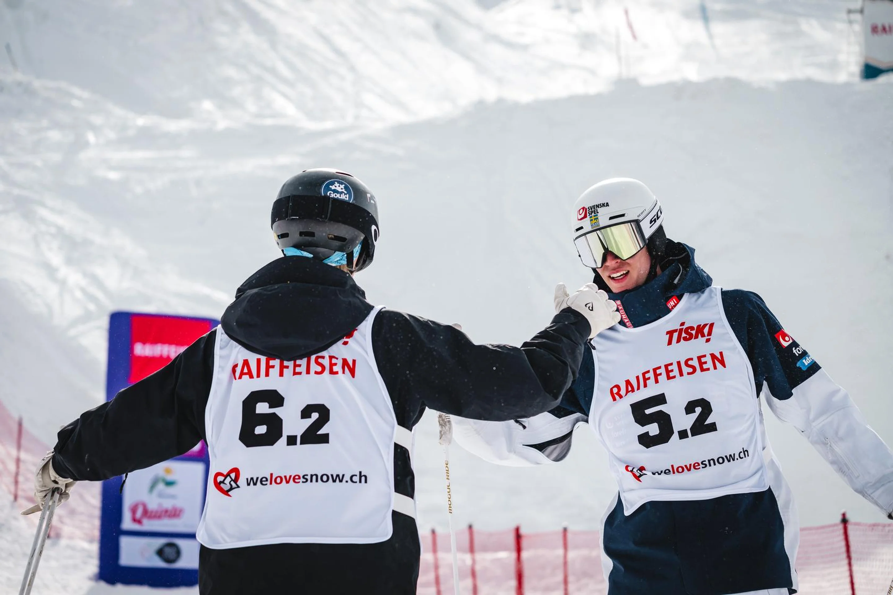 Two skiers wearing helmets and numbered jerseys fist bump on a snowy slope, with red fencing and advertisements in the background.