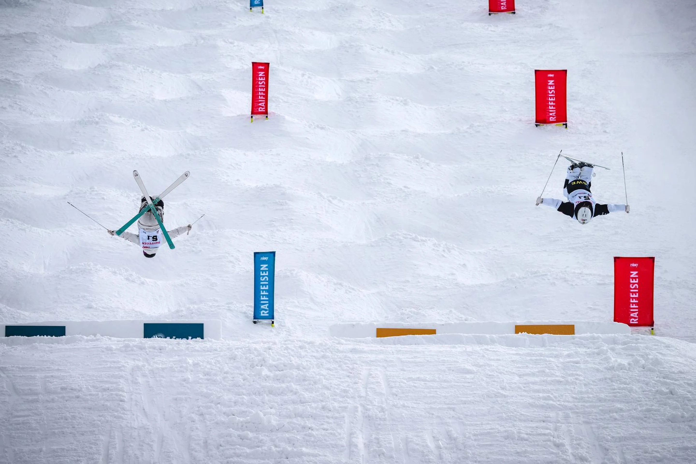 Two skiers perform aerial flips on a snowy slope, surrounded by colored flags marking the course.