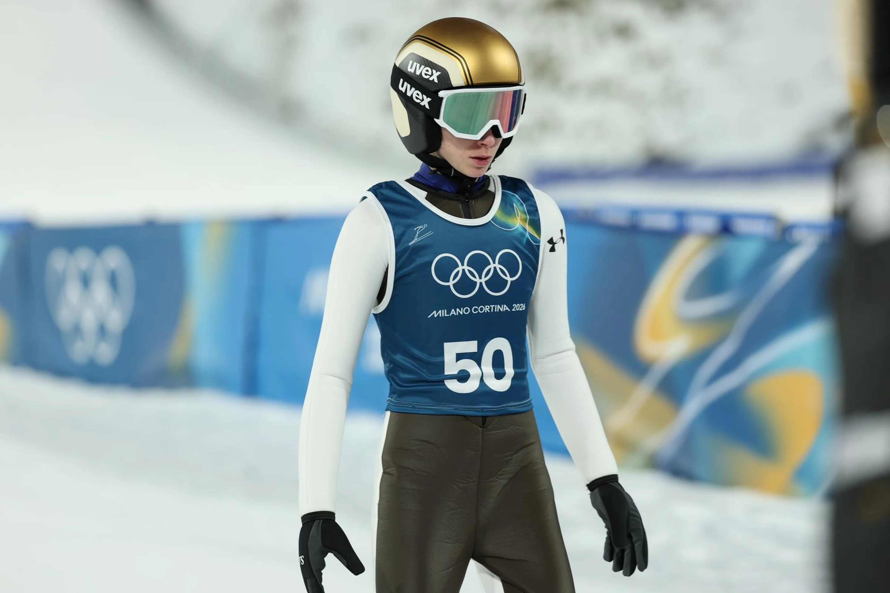 A skier wearing a gold helmet and blue vest with number 50 at a snowy Olympic venue, preparing for a ski event.