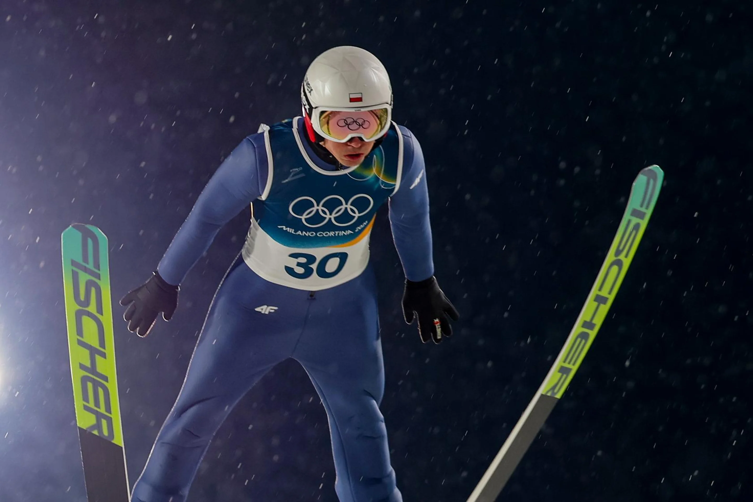 A ski jumper in mid-air, wearing a blue suit and white helmet, with green skis, competing at night surrounded by falling snow.