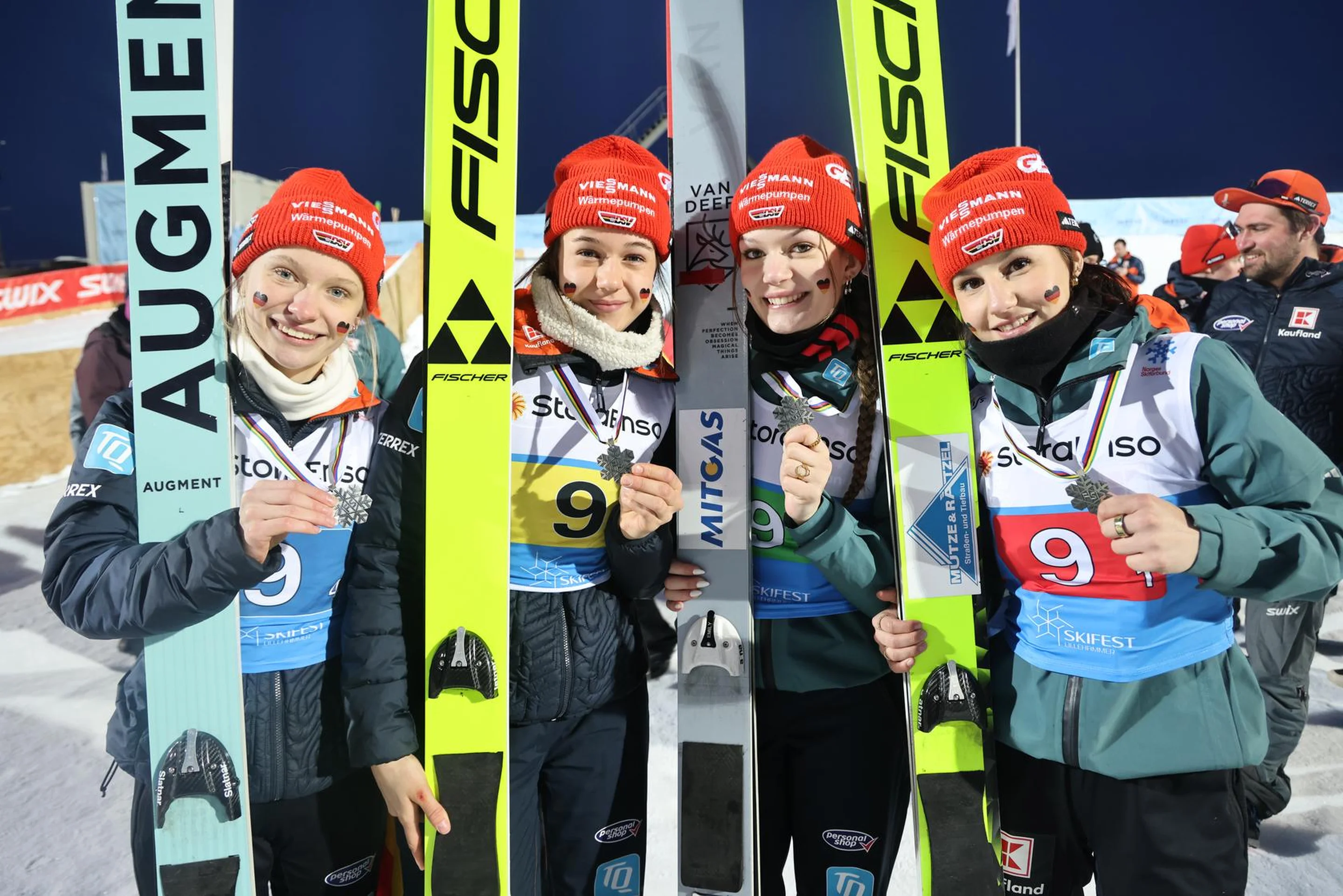 Four ski jumpers wearing red hats and winter gear smile, holding their skis upright, with a snowy background and crowd behind them.