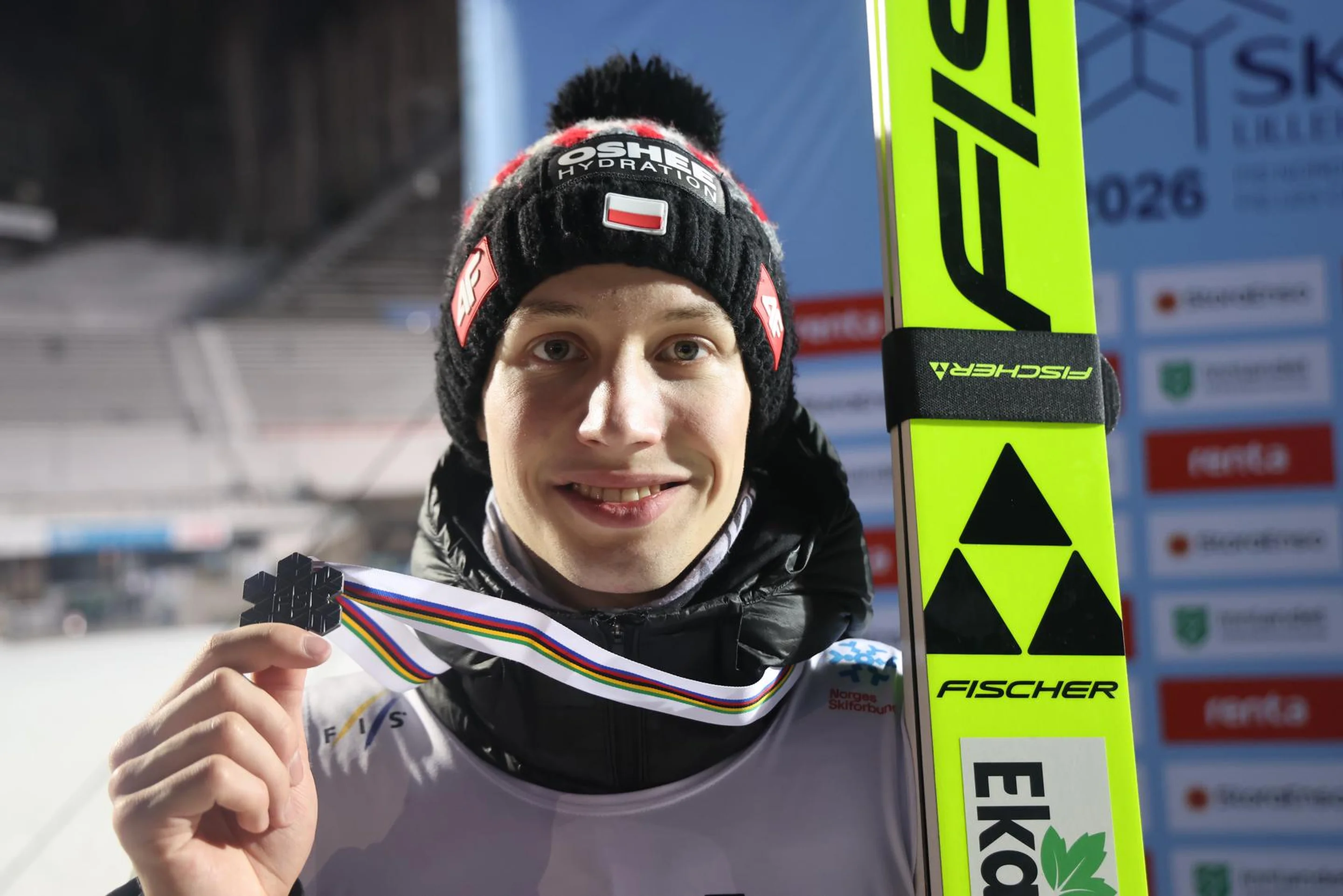 A skier in winter gear smiles while holding a medal in one hand and ski equipment in the other, standing on a snowy ski slope.