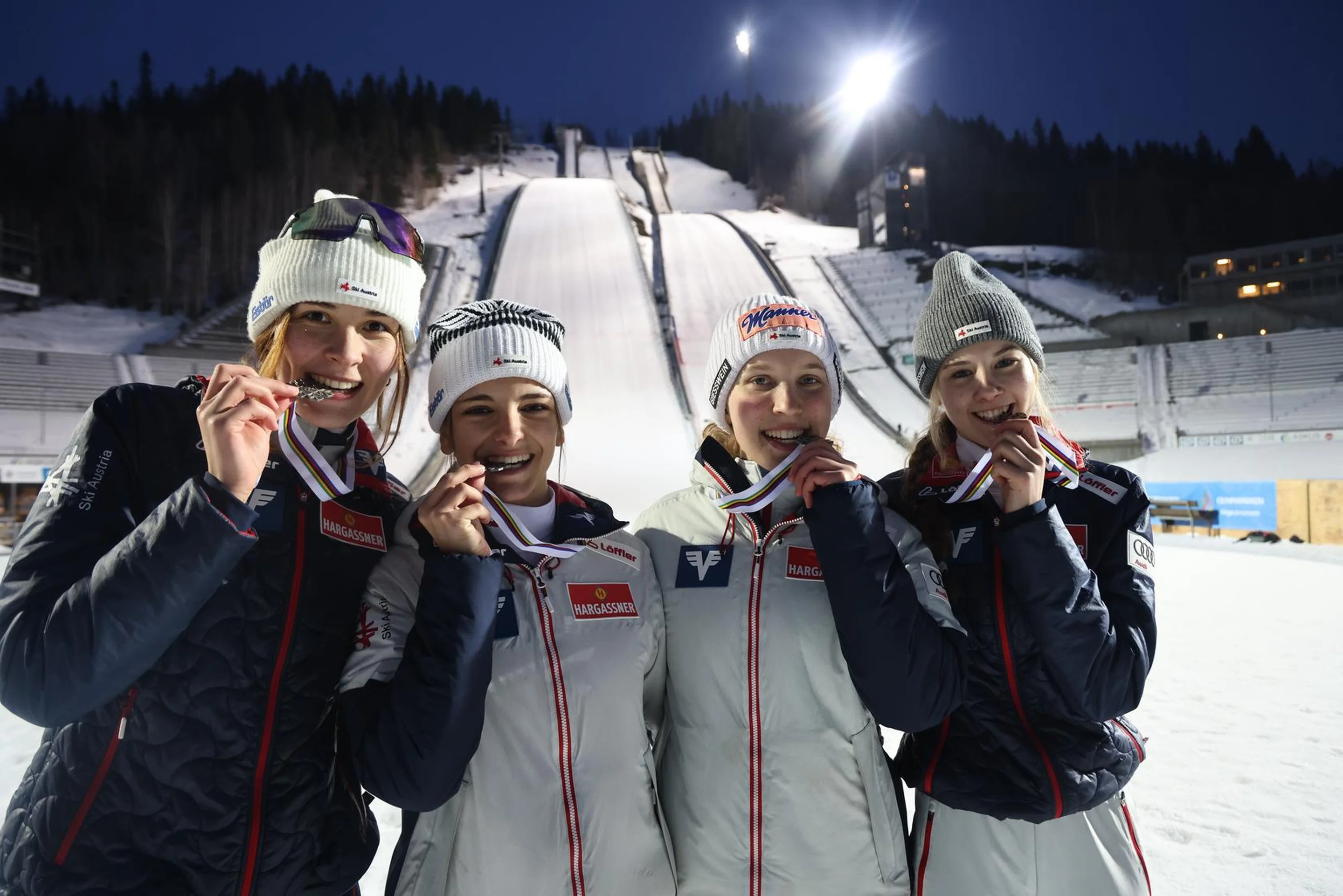 Four athletes in winter gear holding medals, standing in front of a ski jumping hill at night.