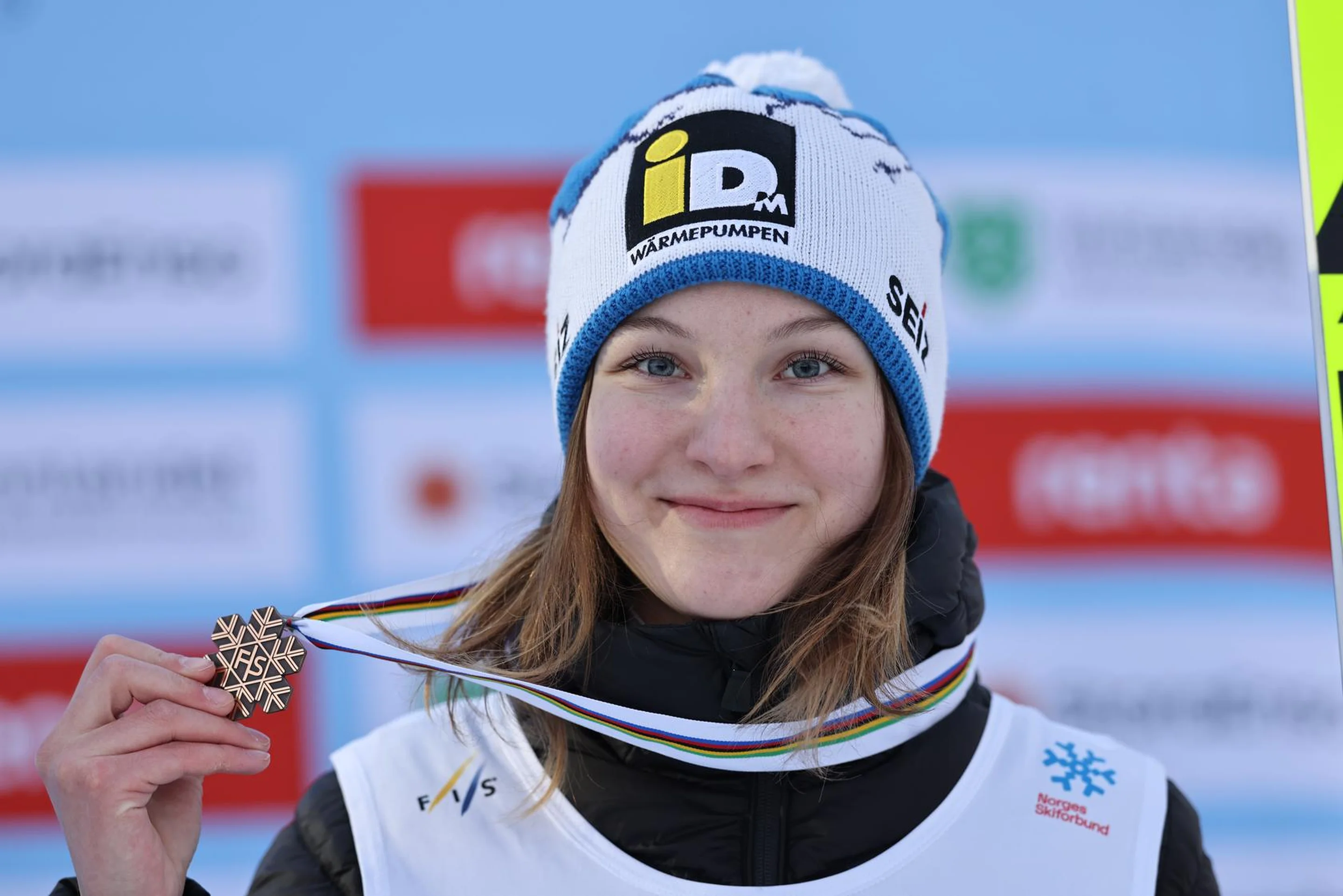 Person smiling, holding a snowflake-shaped medal, wearing a blue hat and white bib, with sponsor logos on a blurred background.