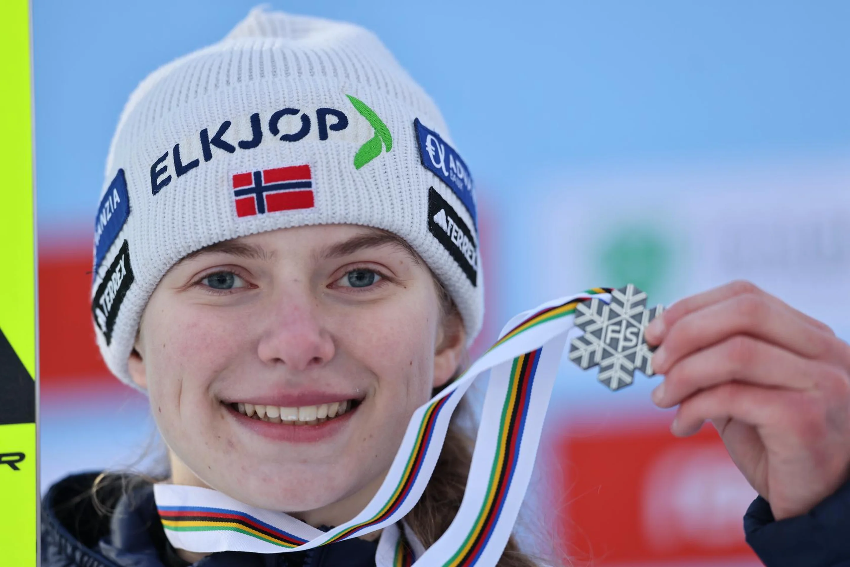 Athlete smiling, holding a silver medal with a snowflake design, wearing a white beanie with logos and a Norwegian flag, set against a blue background.