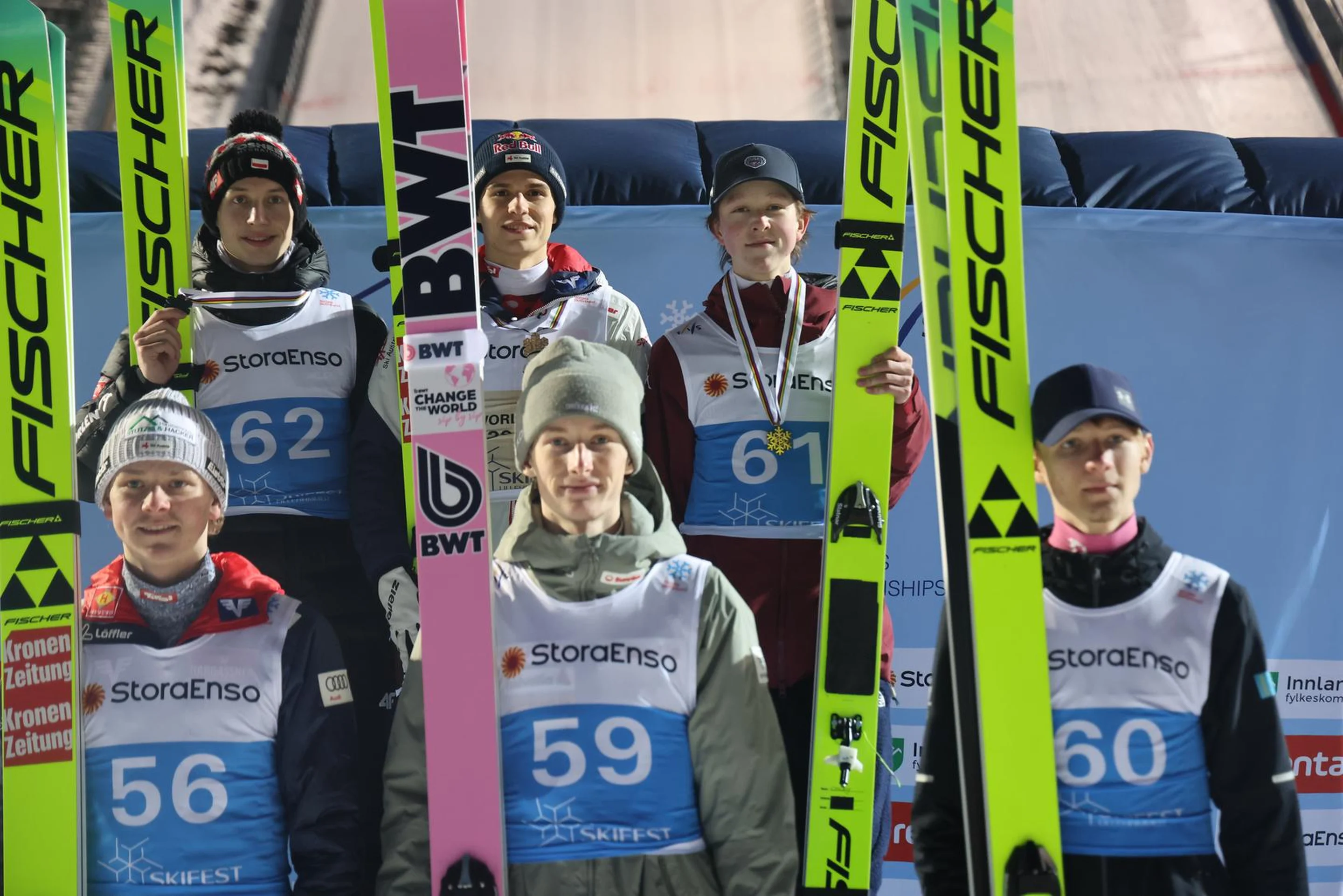Ski jumpers in competition gear holding skis, standing in two rows on a podium in front of a ski jumping ramp.