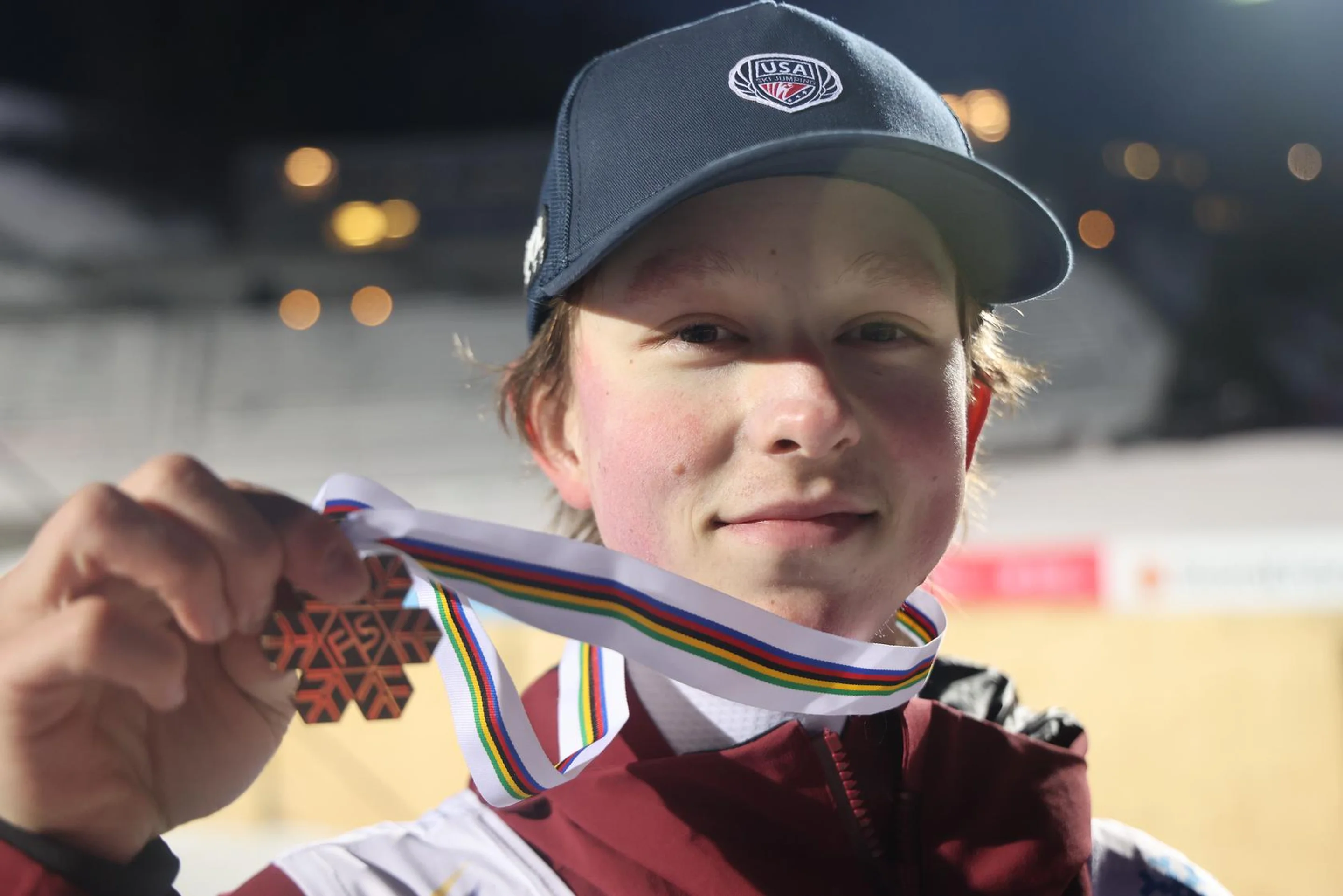 Person wearing a cap holds up a sports medal with a colorful ribbon, smiling slightly, with blurred background.