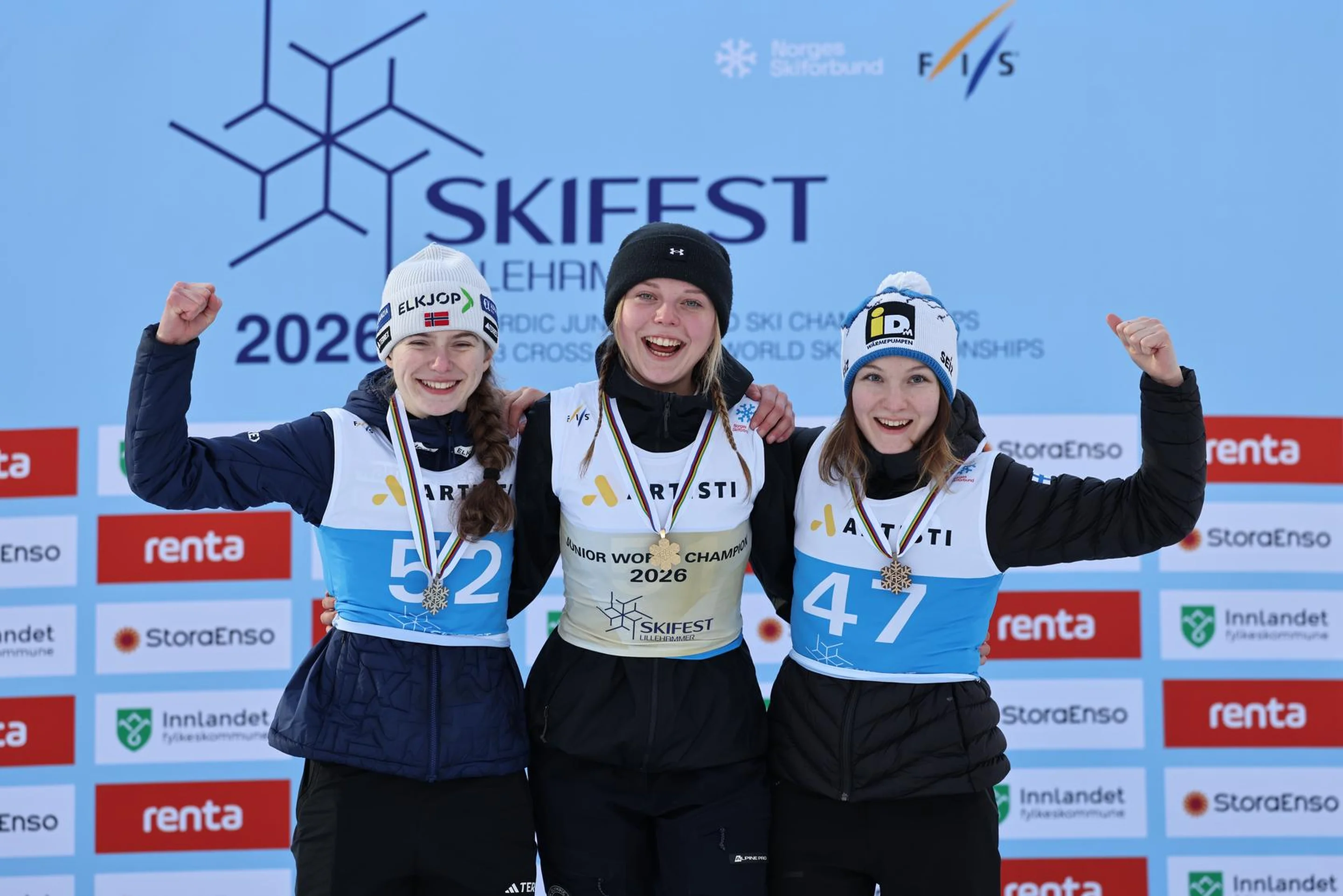 Three smiling skiers in winter gear celebrate on a podium against the Skifest 2026 backdrop, each wearing numbered bibs and medals.