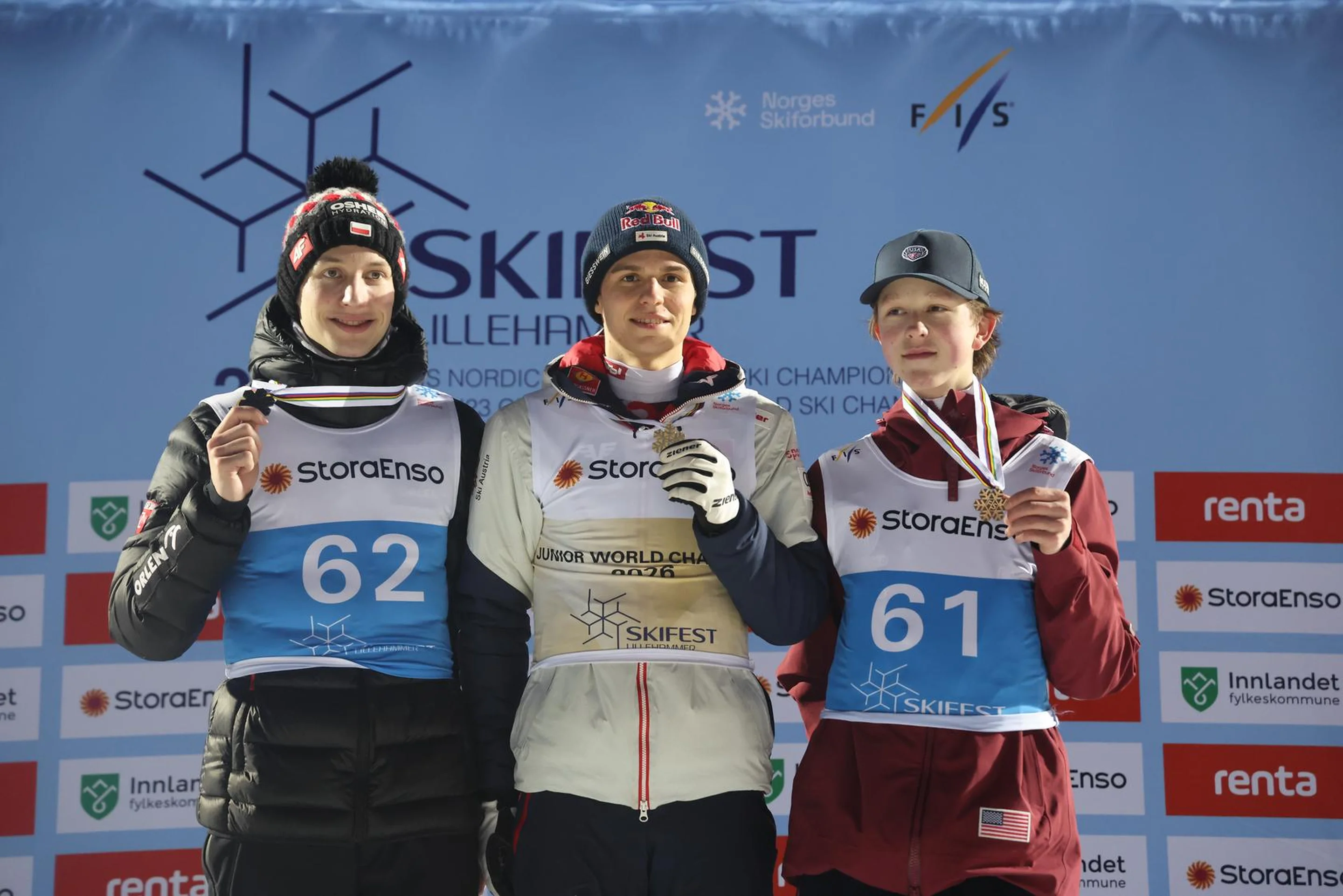 Three skiers stand on a podium with medals, smiling. They wear numbered bibs and winter gear, with a Skifest backdrop behind them.