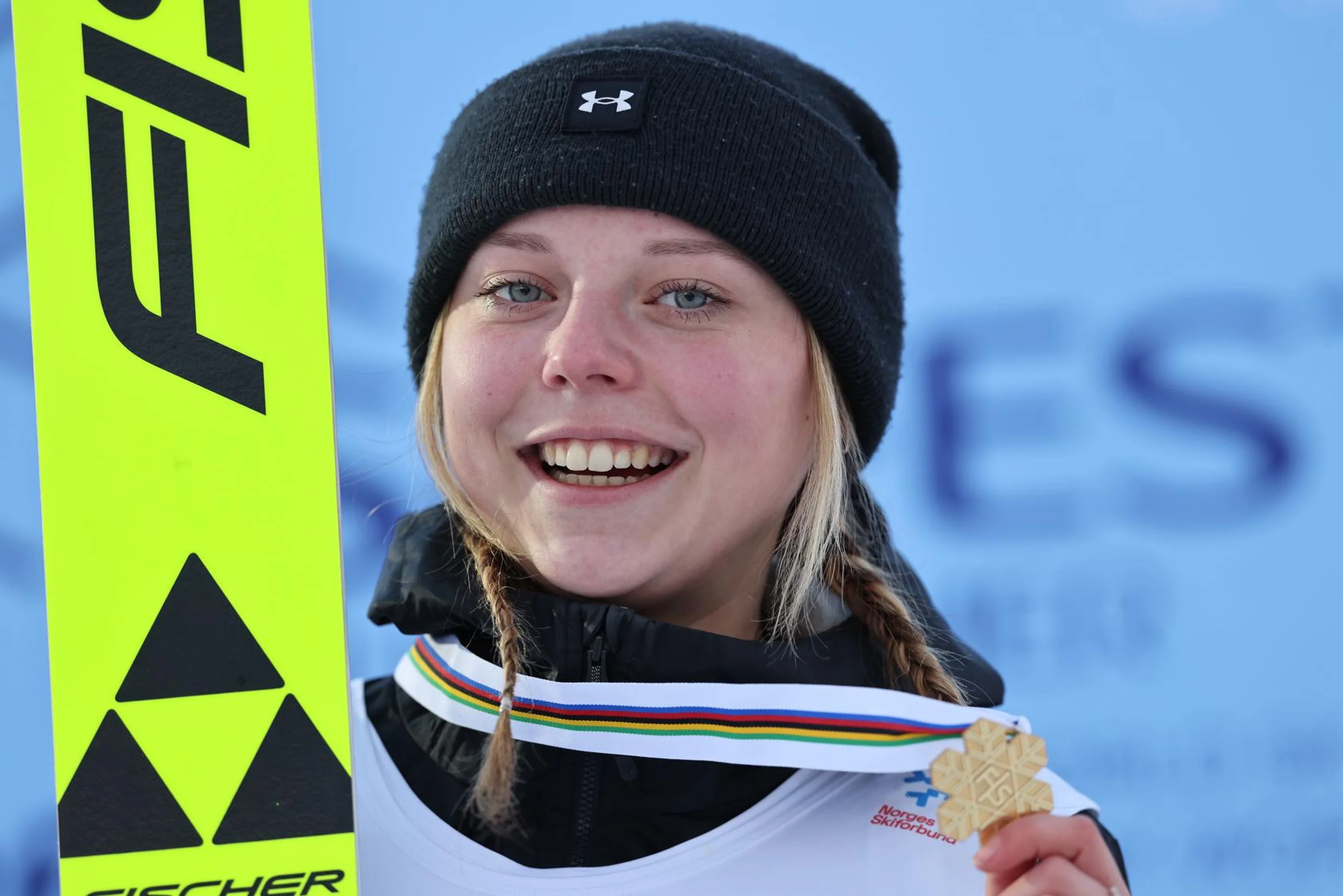 Smiling athlete with a black beanie holds a gold medal and ski in front of a blue sponsor backdrop.