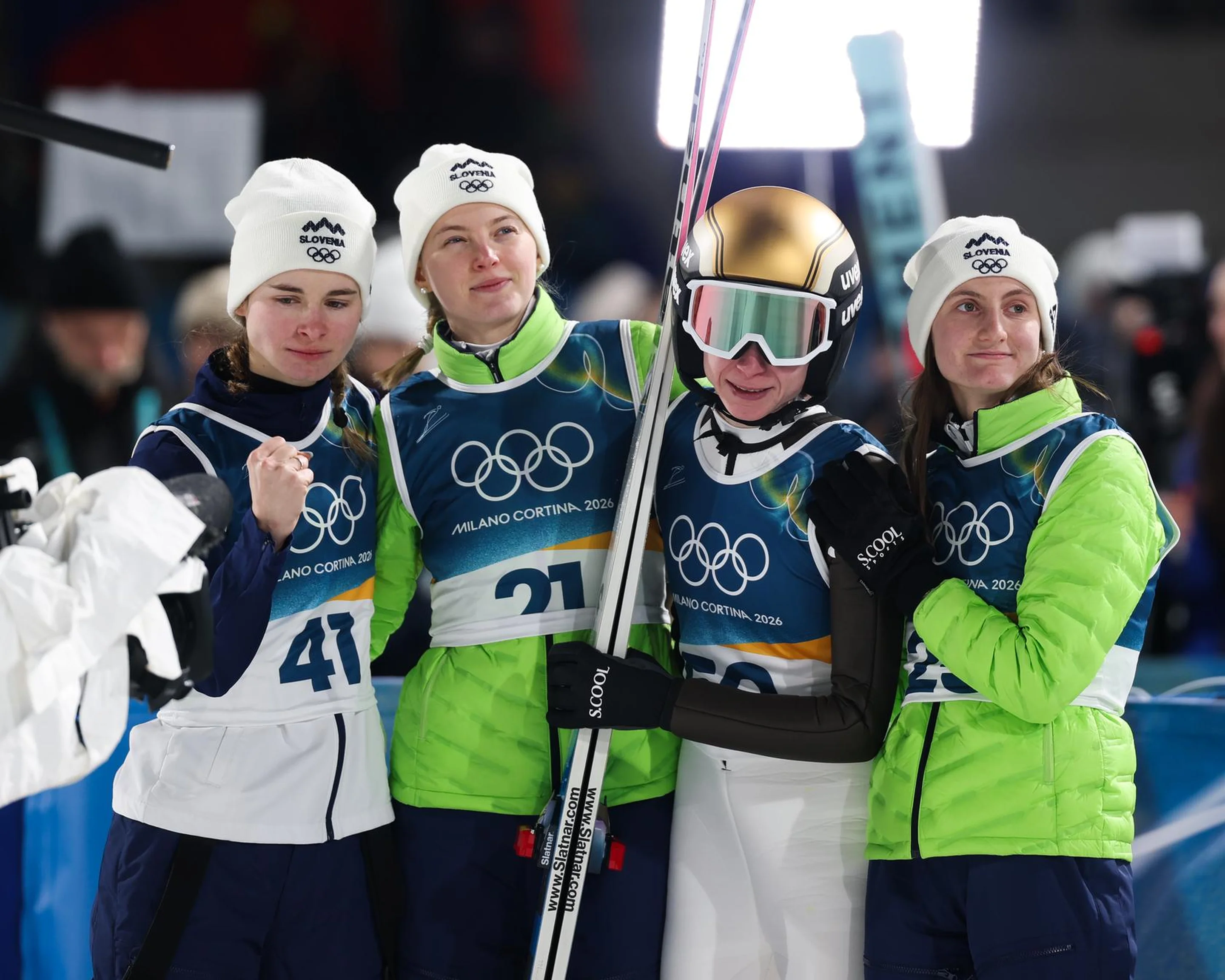 Four ski athletes in green and white uniforms and helmets stand together, wearing Olympic badges, holding skis, and smiling for a photo.