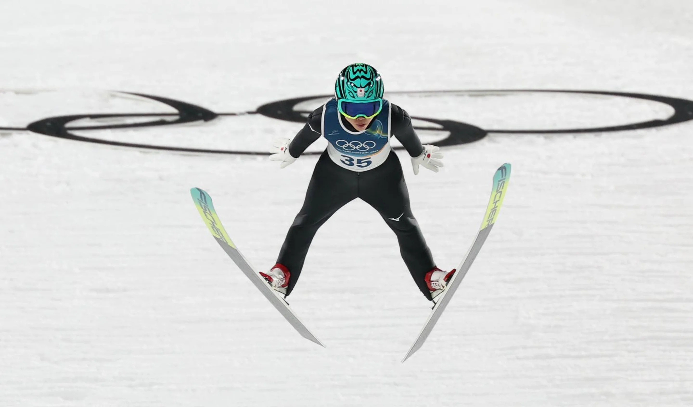 Ski jumper in mid-air with skis apart, wearing green helmet and goggles, Olympic rings visible in snow background.