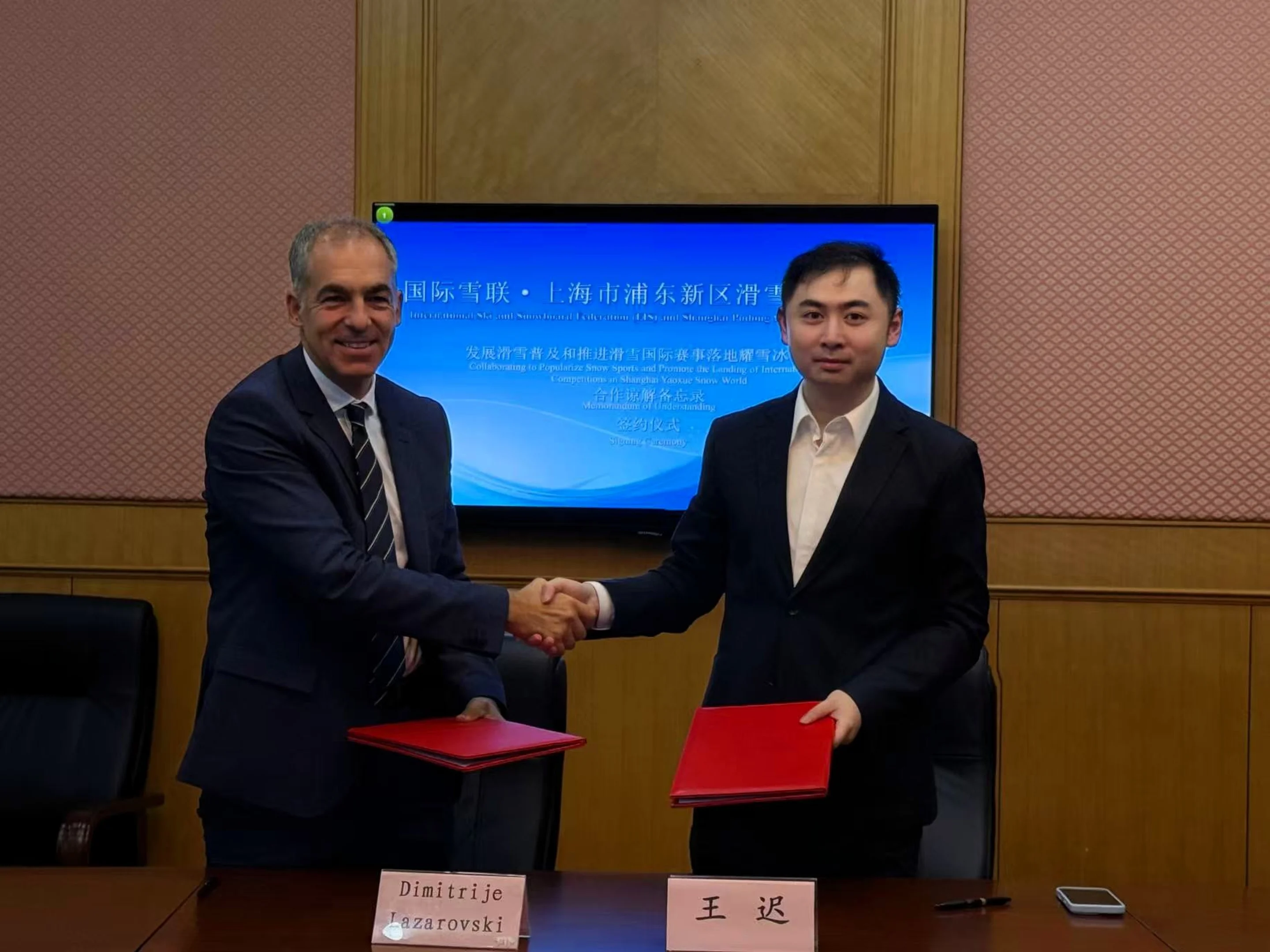 Two men in suits shaking hands, holding red folders, with a presentation screen in the background. Nameplates are on the table.