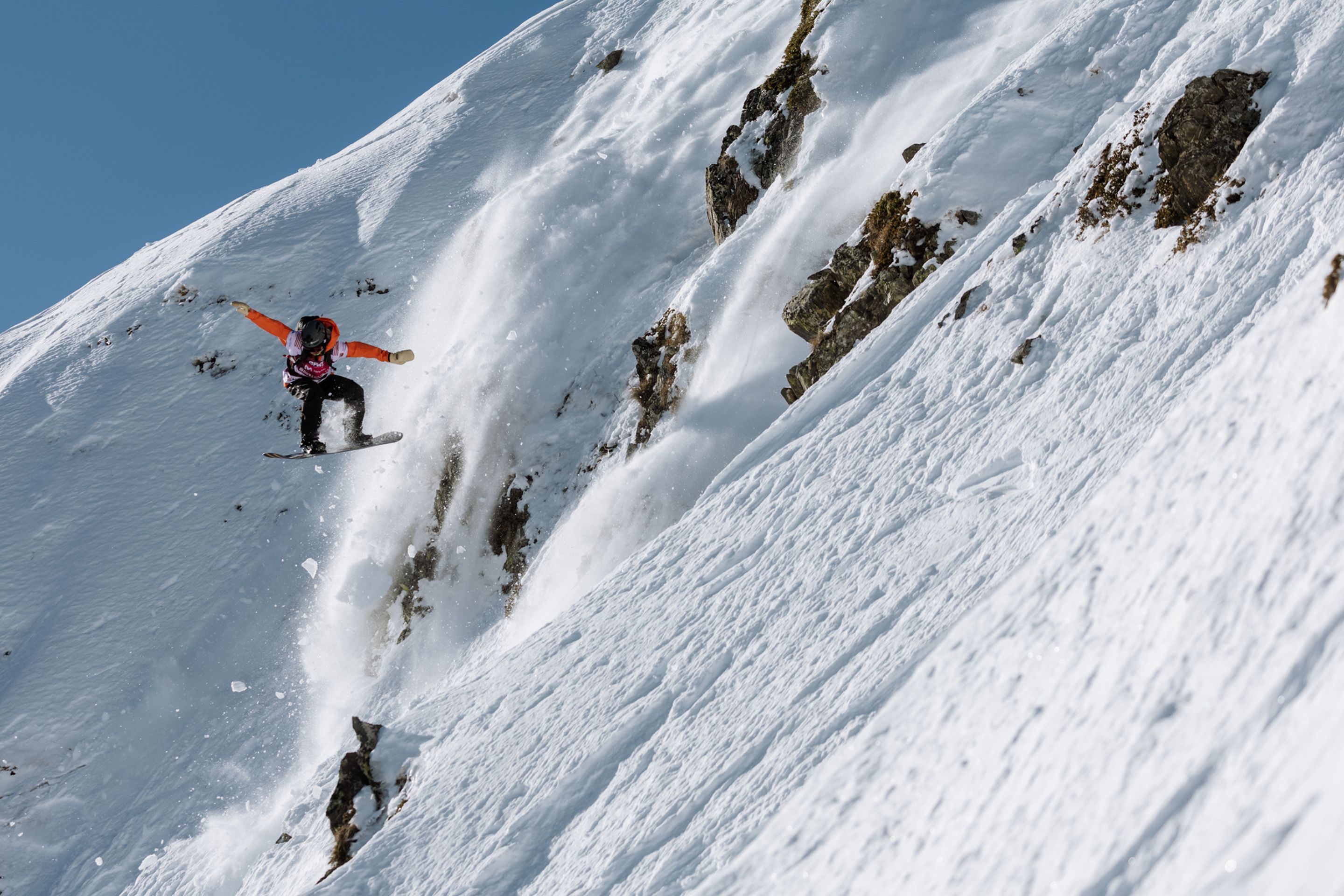 A snowboarder in mid-air descends a steep, snowy mountain slope with rocky patches under a clear blue sky.