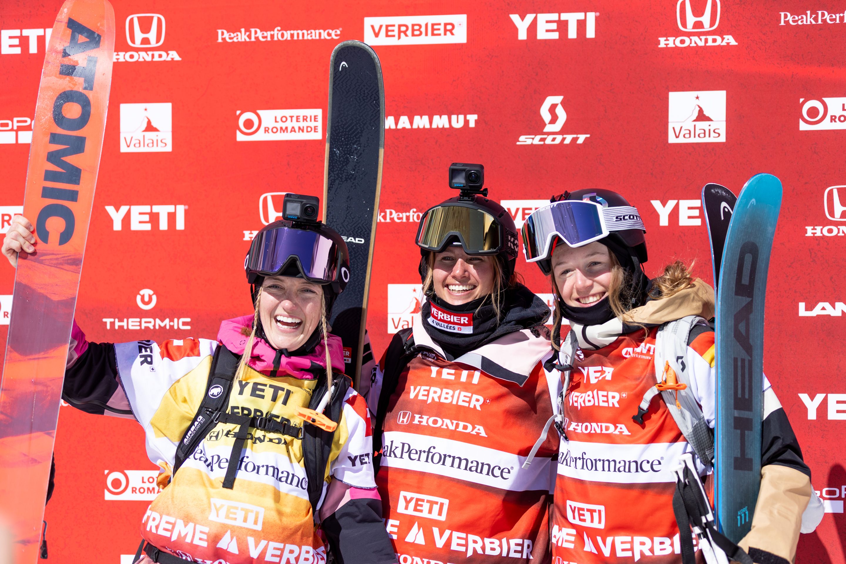 Three smiling skiers in colorful gear pose with skis against a red sponsor backdrop, celebrating at a winter sports event.