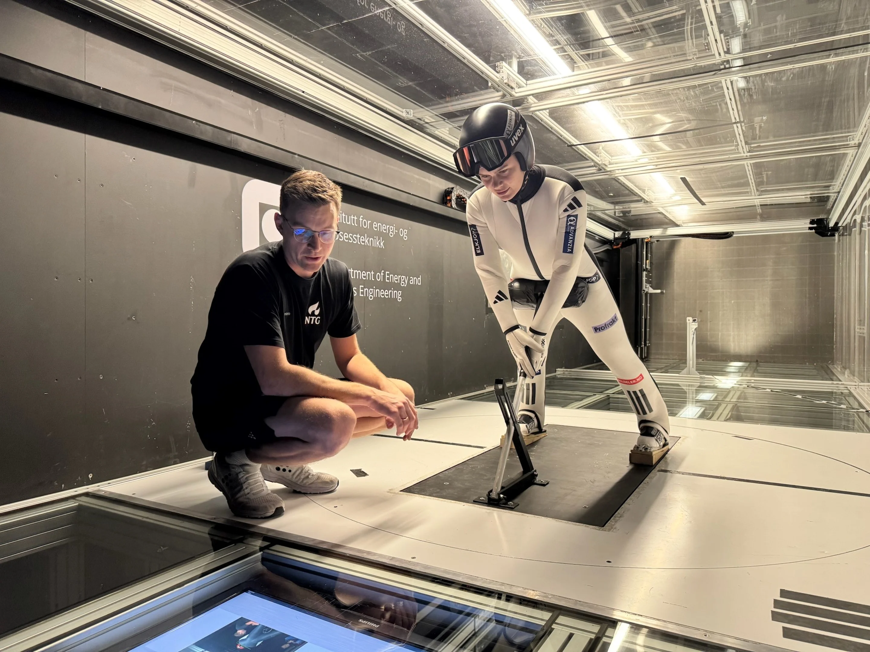 A person crouches next to a ski jumper in a wind tunnel, both focused on testing equipment. The ski jumper wears a white suit and helmet.