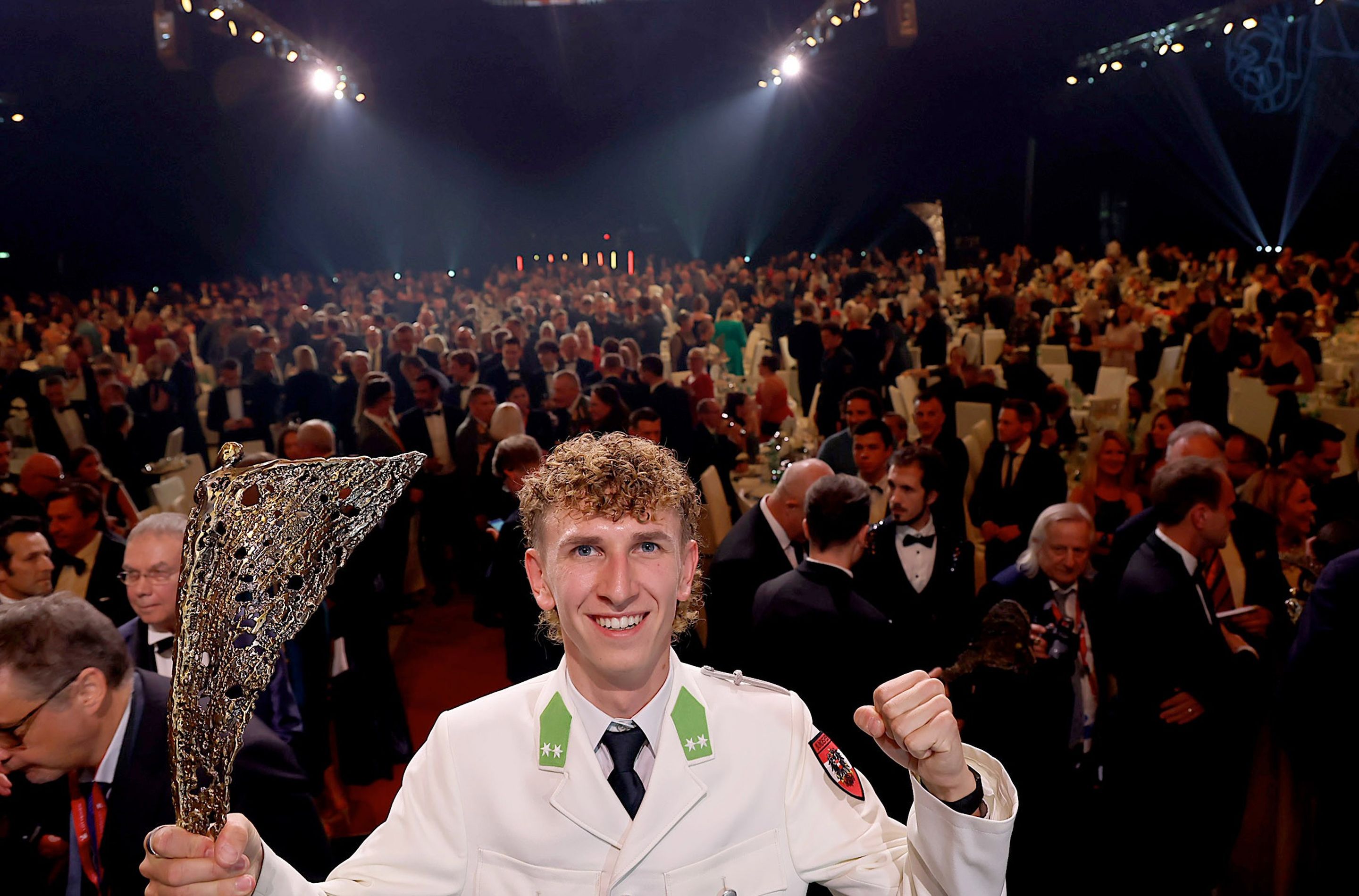 A person in a white uniform holds a trophy, smiling, with a large, seated crowd in formal attire in the background.