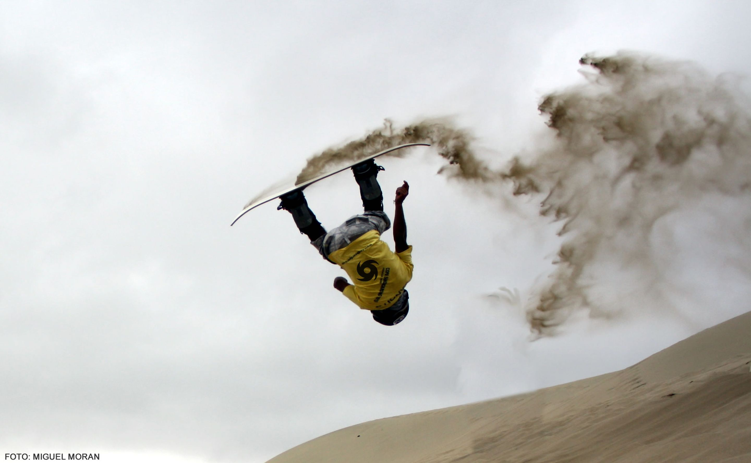 Victor-Segundo Chavez, in a yellow t-shirt, performs a backflip while sandboarding, sand flying off his board