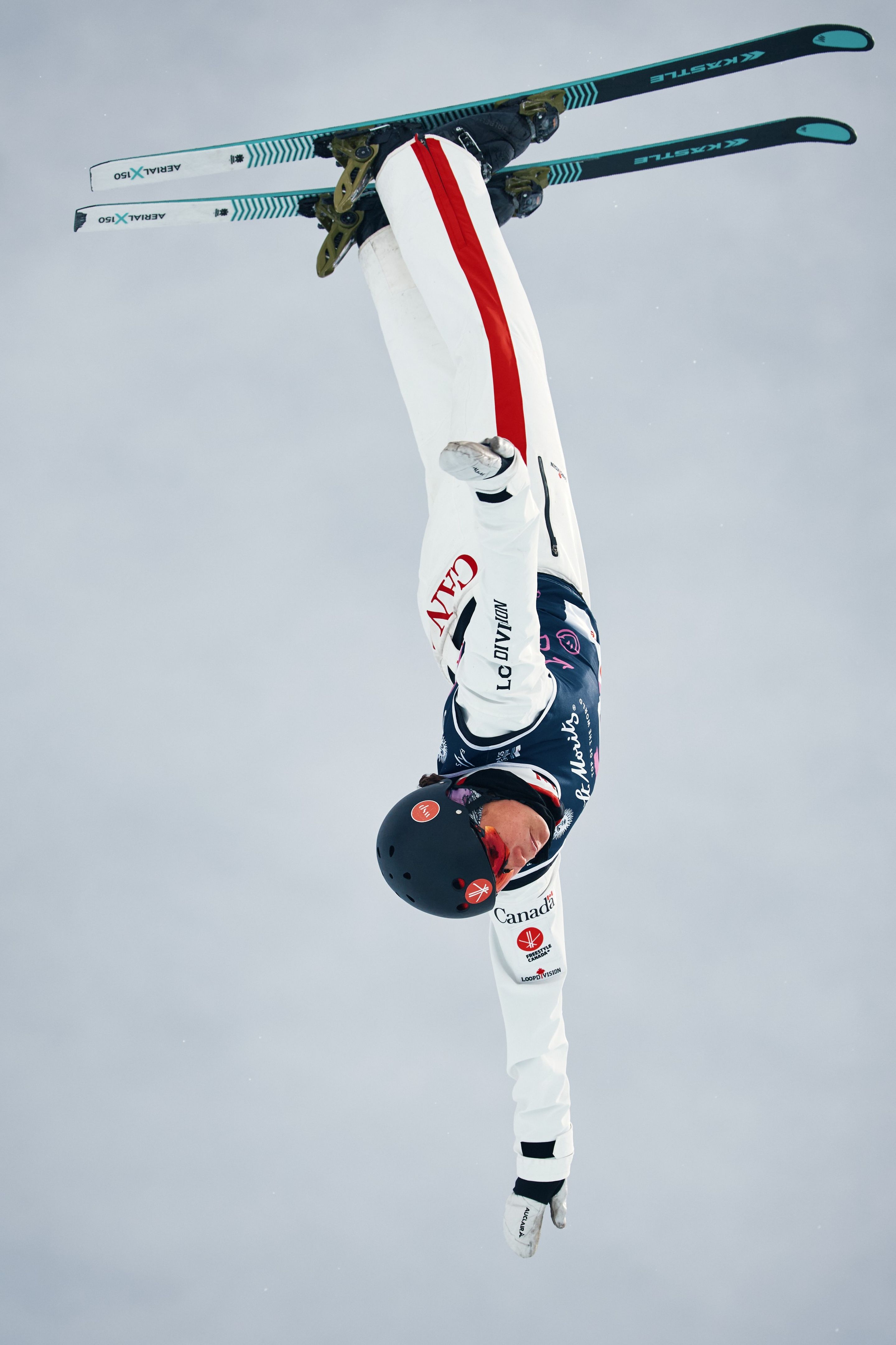 A skier performs an inverted aerial trick against a snowy backdrop, wearing a white and black outfit with red accents.