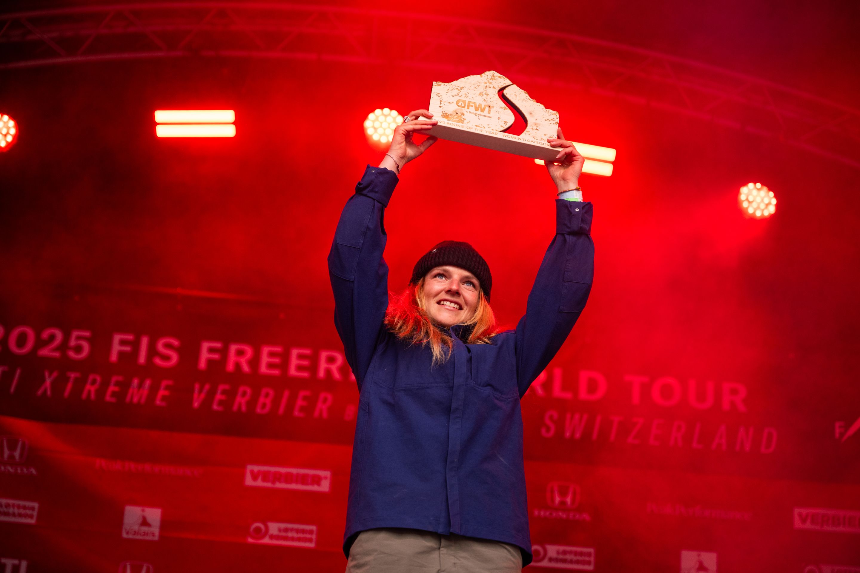 Person in a blue jacket and beanie holds up a trophy triumphantly on stage, with red lighting and event banners in the background.
