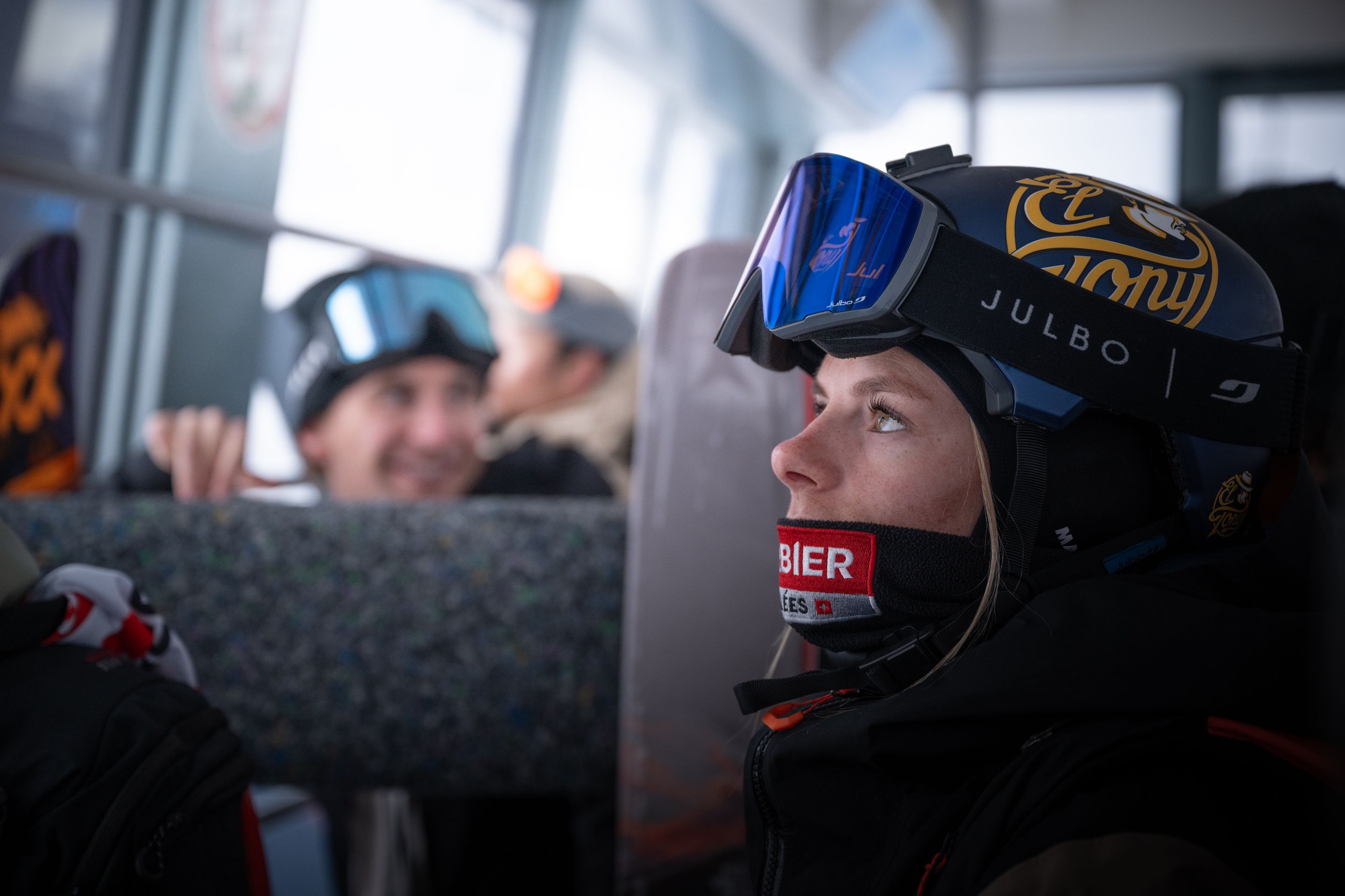 A person in ski gear and goggles looks out a window in a ski lift, with others seated nearby.